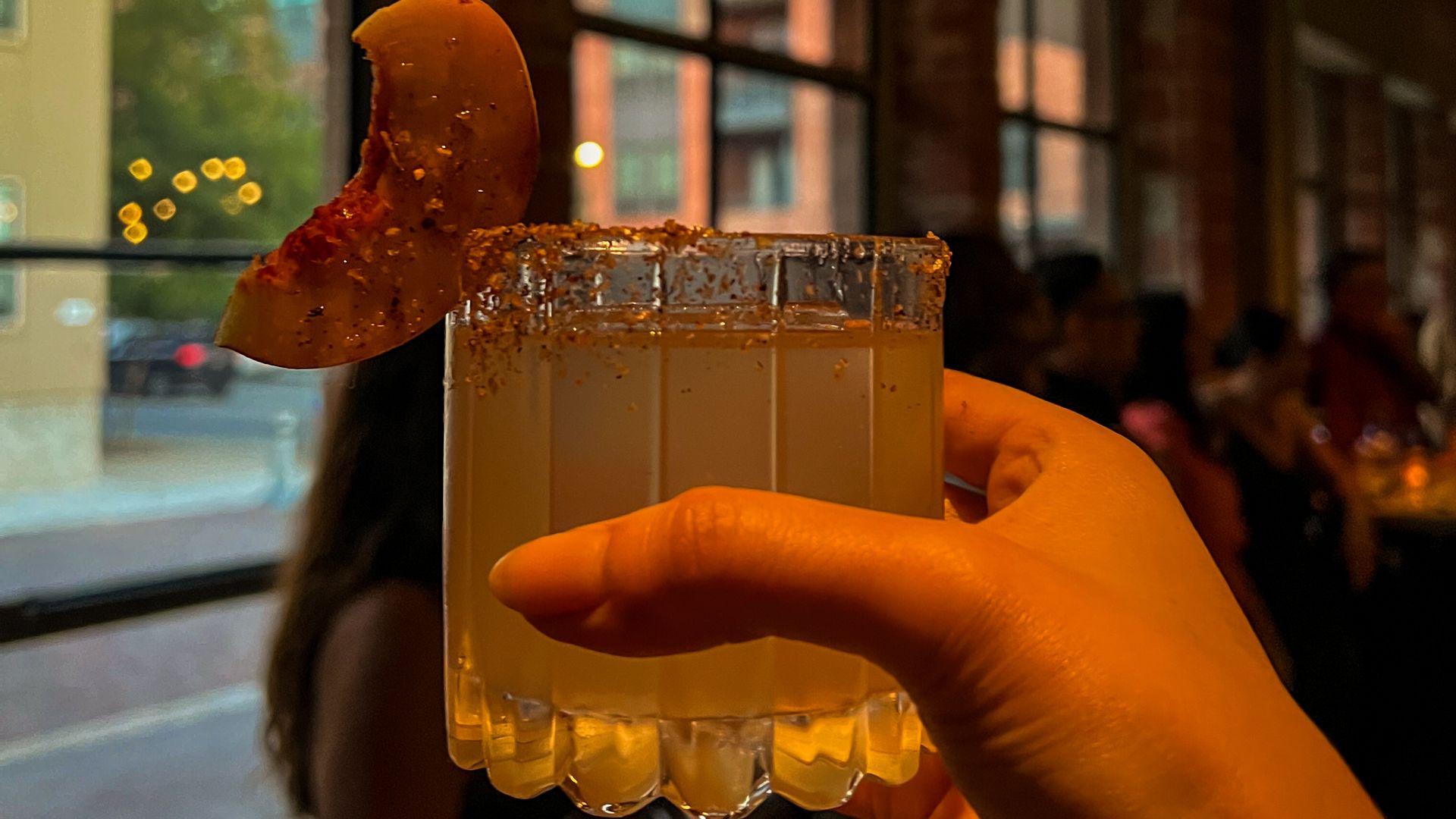 A hand holding a clear cocktail glass , garnished with a peach slice and filled with a light-colored liquid.