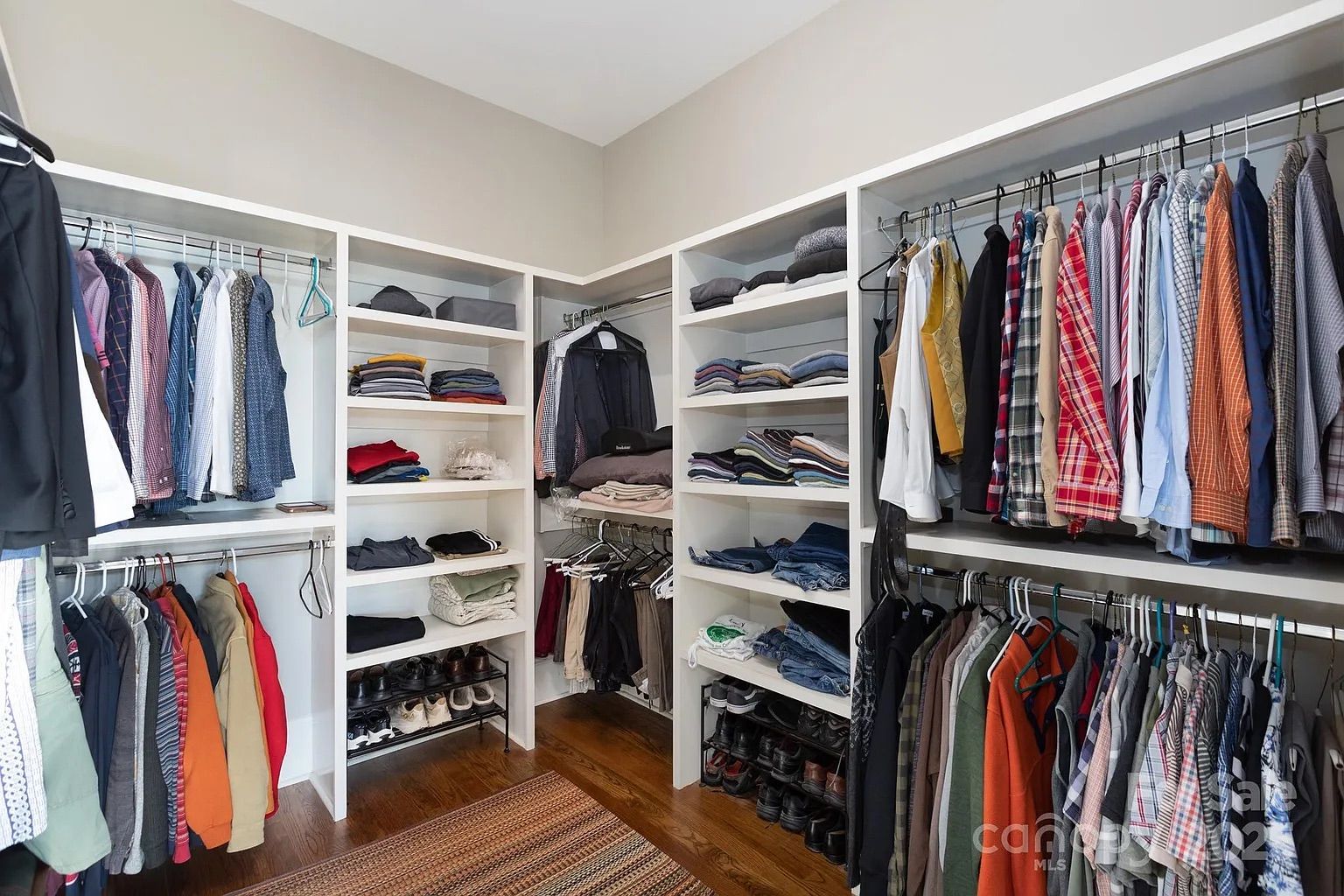 Neat walk-in closet with white shelves and rods holding folded clothes, shoes, and a variety of colorful shirts, jackets, and pants on hangers against a beige wall and wooden floor.