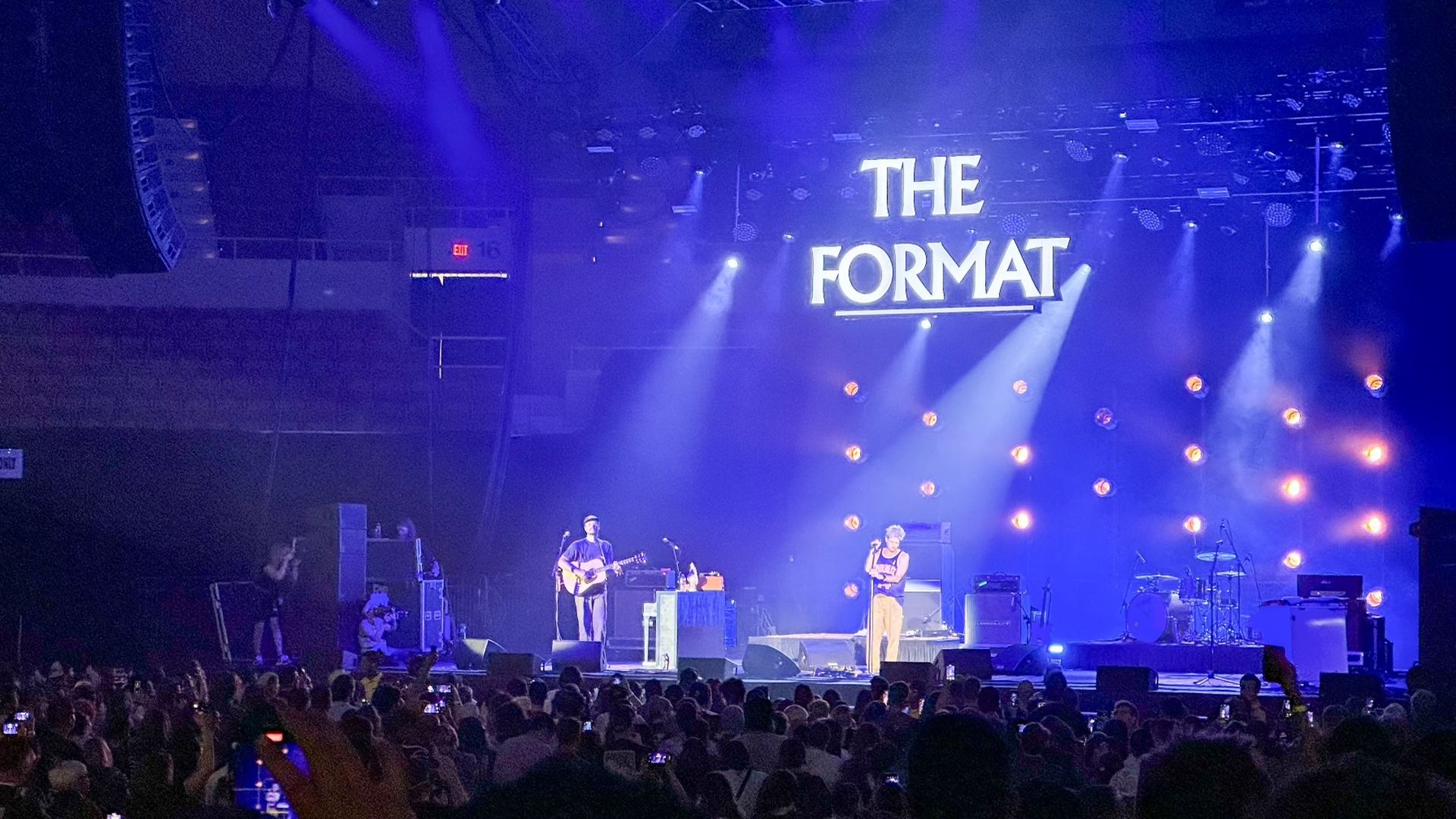 Concert stage with blue and purple lights and a crowd. Two performers stand under a large sign reading "THE FORMAT" amid bright circular stage lights.