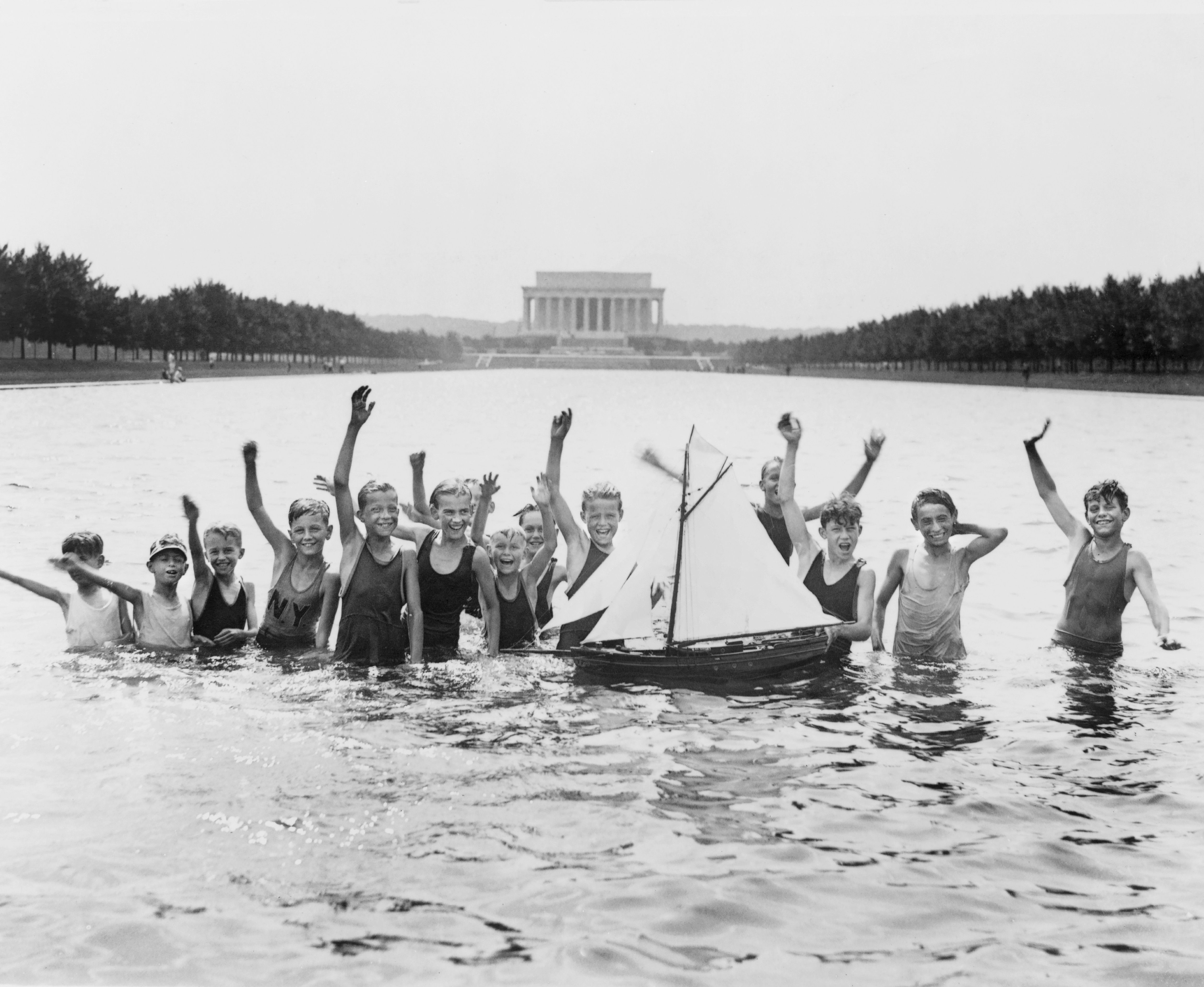 A group of boys waving at camera while Playing with a toy sailboat in the Reflecting Pool in front of the Lincoln Memorial, Washington DC, July 7, 1926 . 