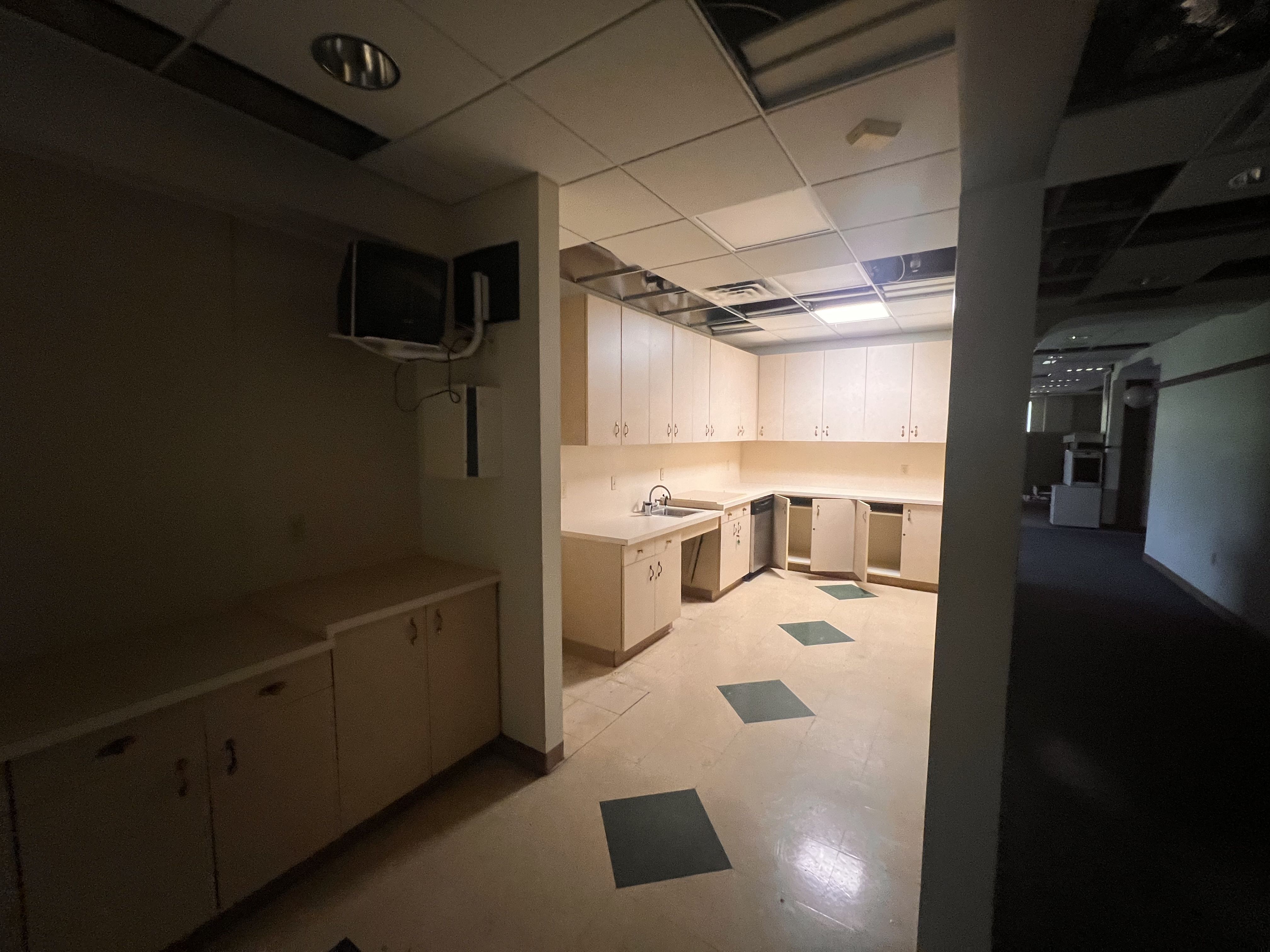 Dim office kitchenette with beige cabinets along a back wall, a sink, and a wall-mounted TV on the left. Damaged ceiling tiles, a dark hallway, and a pale pillar frame the space.