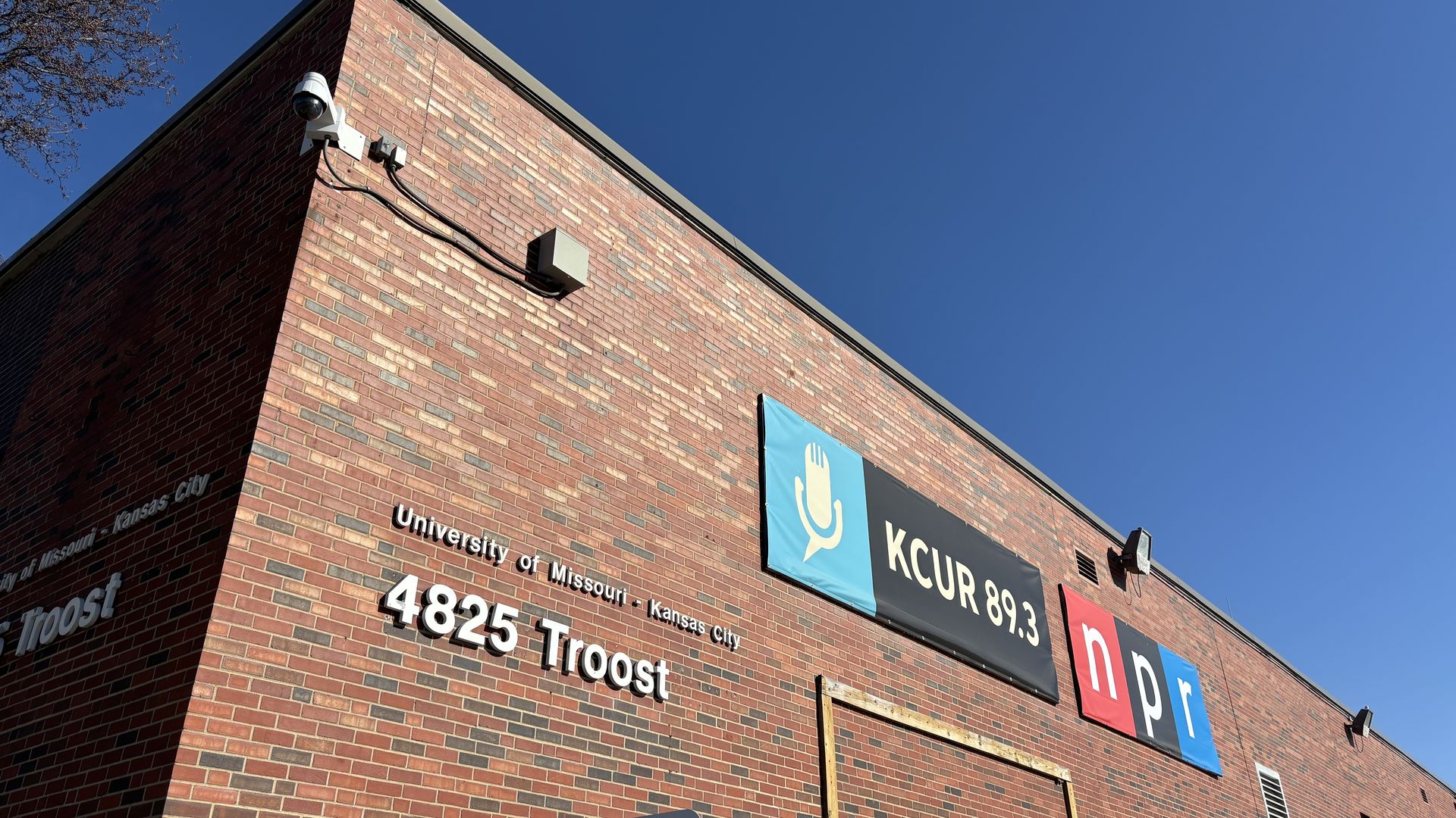 Red brick building wall with signs for KCUR 89.3 and NPR, located at 4825 Troost, University of Missouri - Kansas City, under a clear blue sky.