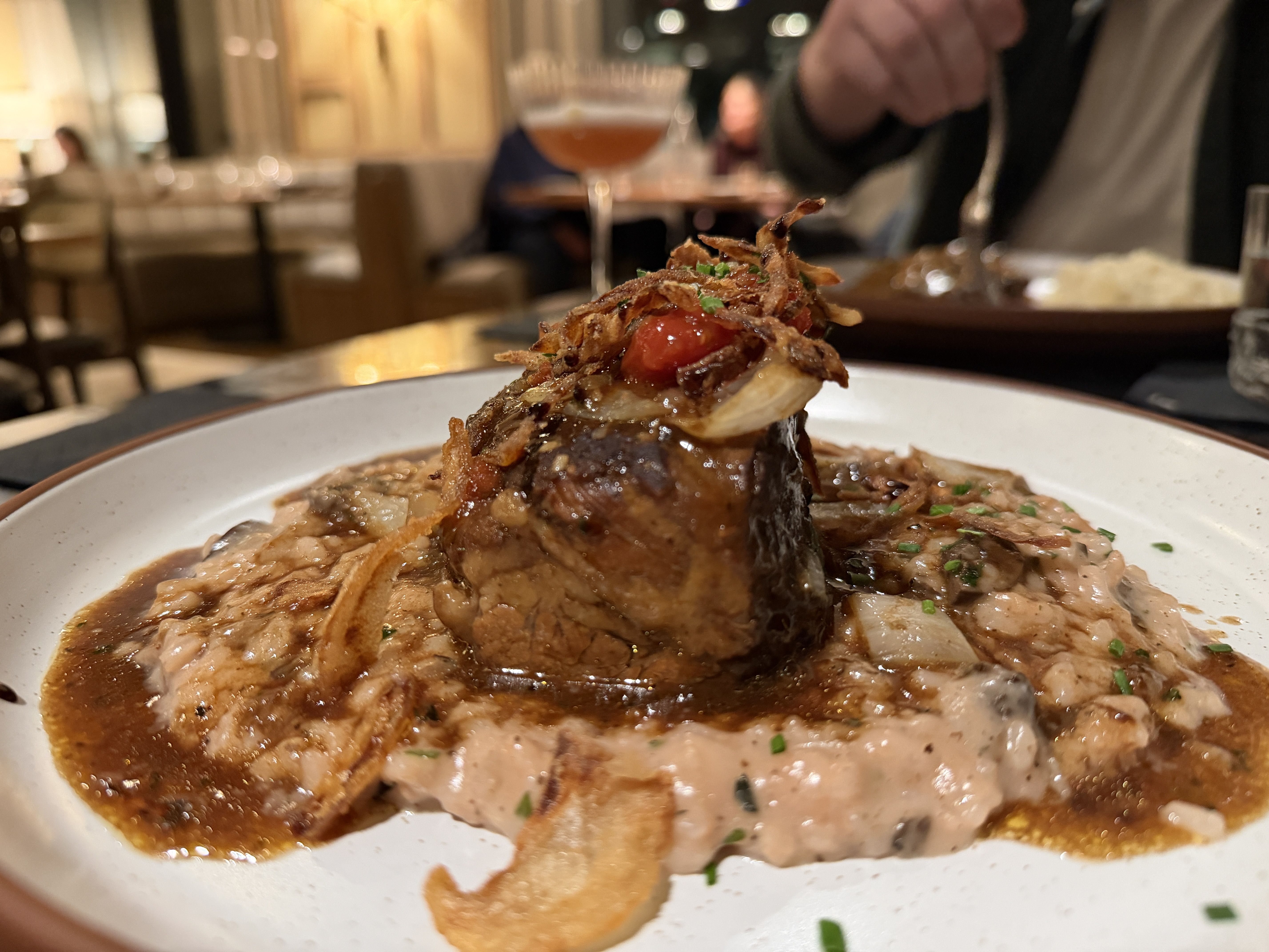 Close-up of a plated gourmet dish featuring a seared piece of meat topped with crispy fried onions and a red garnish, served on a creamy mushroom risotto with a rich brown sauce, in a warmly lit restaurant.