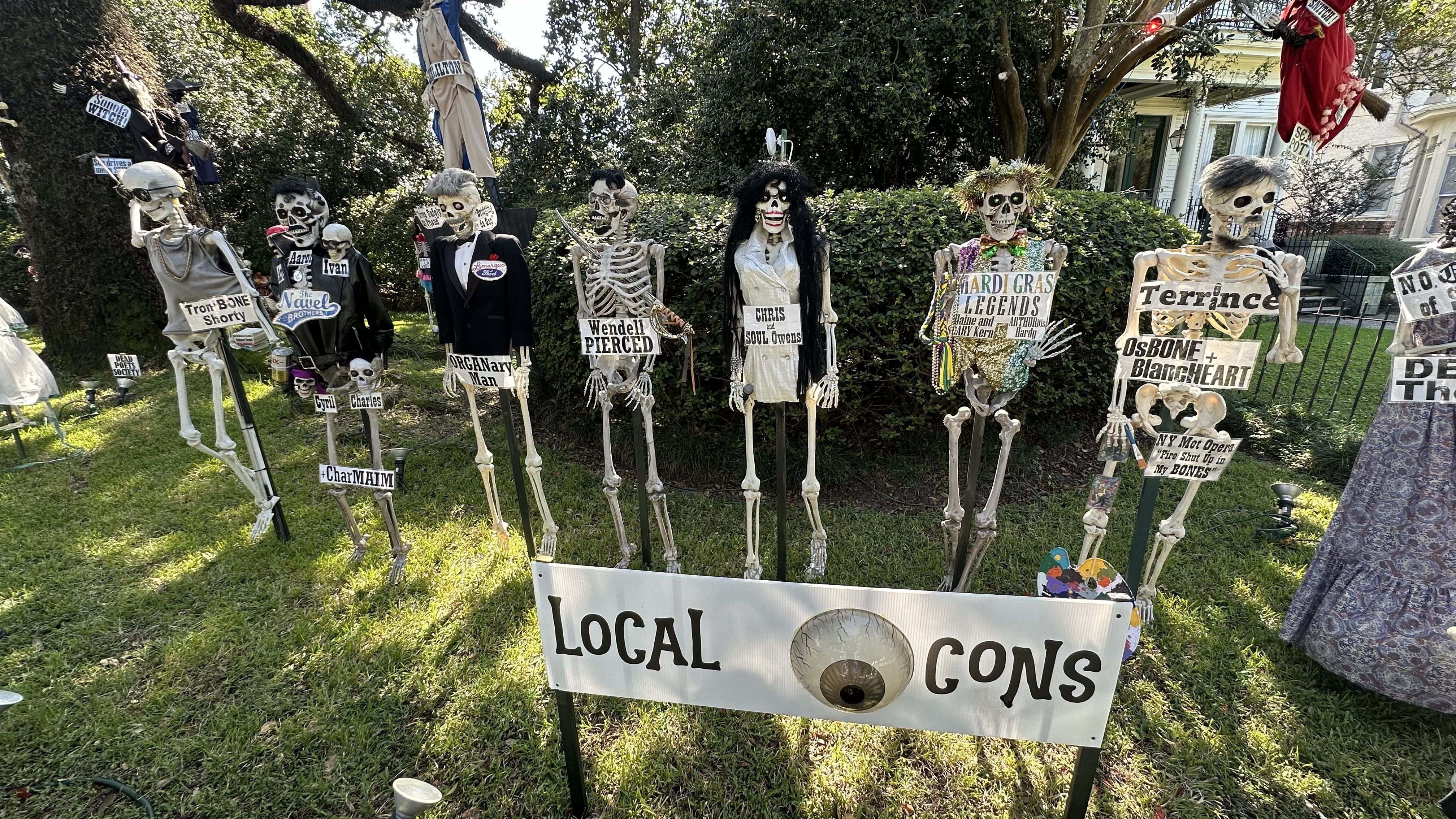 Skeletons decorate a yard and have signs with puns as "local icons." 