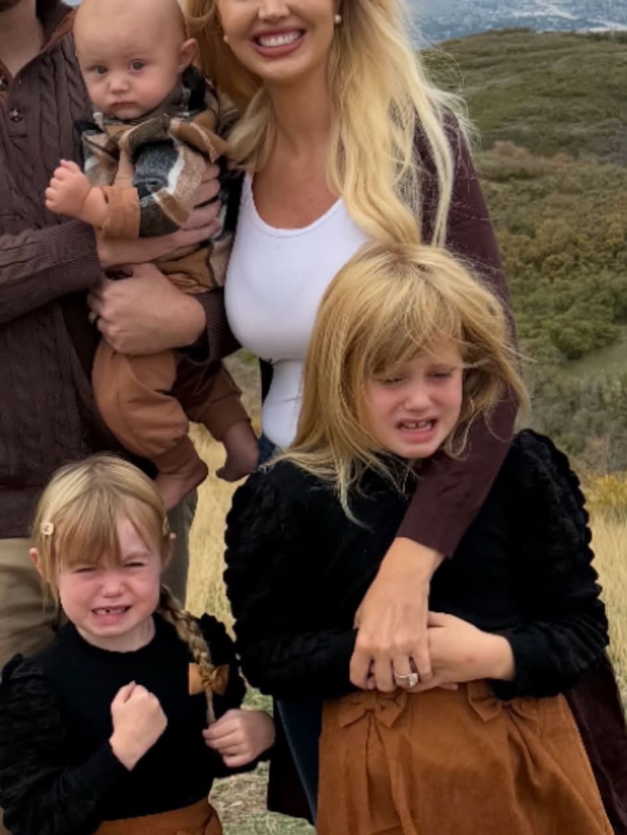 Blonde woman in a white top stands beside a man holding a baby in brown plaid; two girls in black tops and brown skirts pose on a grassy hillside.