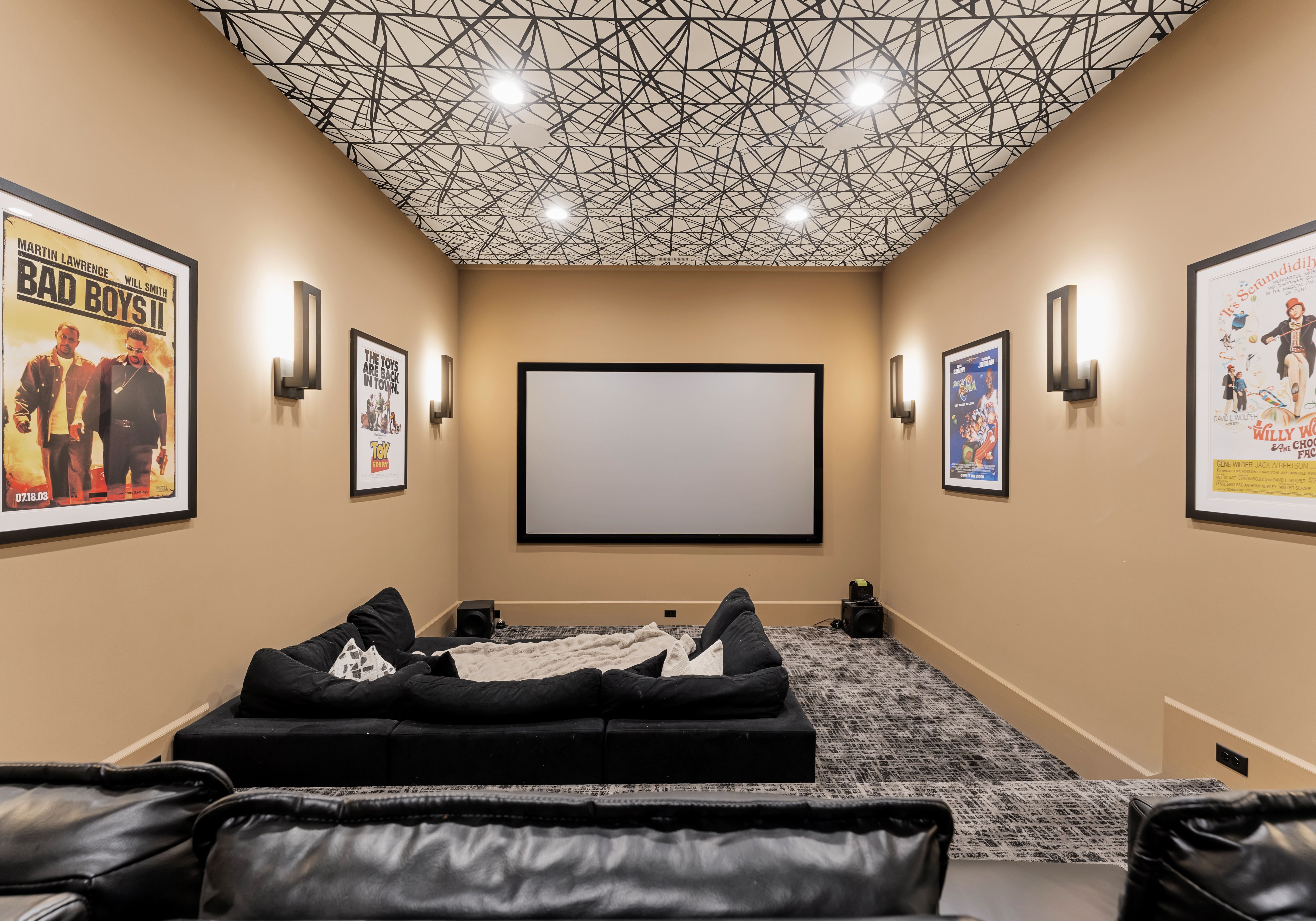 Home theater room with beige walls, patterned black and white ceiling, large black sectional sofa, movie posters including "Bad Boys II," "Toy Story," and blank projector screen.