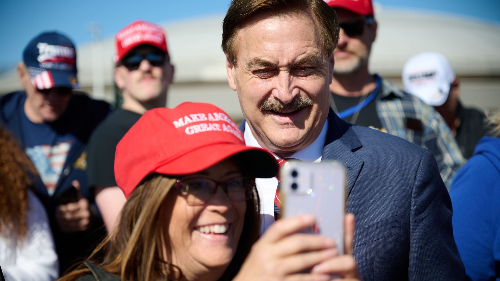 Close-up of a man in a blue suit smiling as supporters surround him; a red cap reads "Make America Great Again" and fans take selfies on a sunny day.