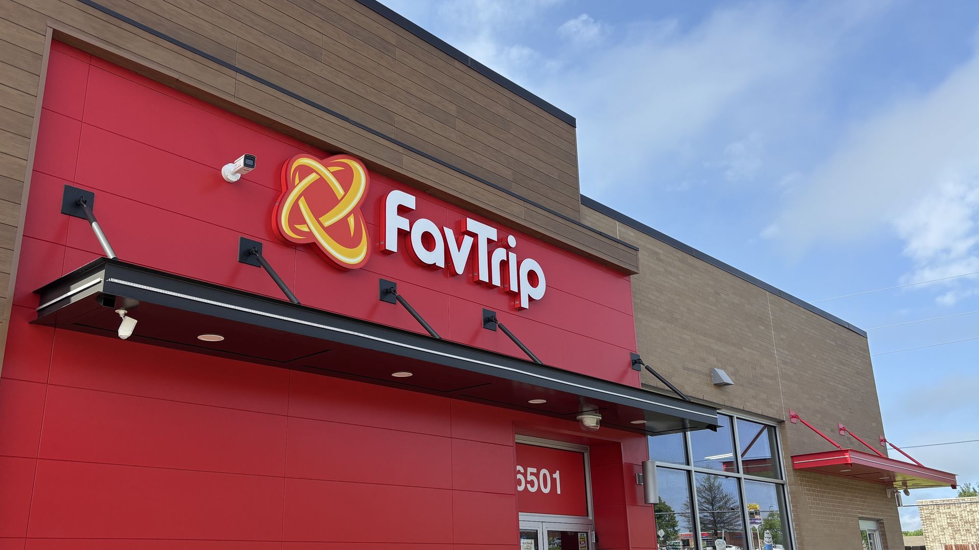 Modern storefront with a red exterior panel and a large illuminated sign that reads "FavTrip" next to a yellow intertwined ring logo; tan brick above, security camera, and a blue sky.