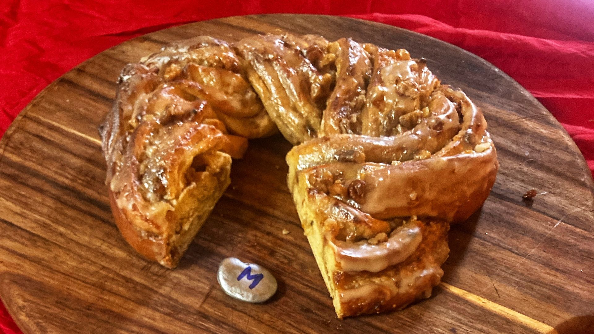 Glazed cinnamon king cake with pecans on a wooden board, one slice placed on a cream-colored plate with decorative edges and a fork, all on a red cloth background.