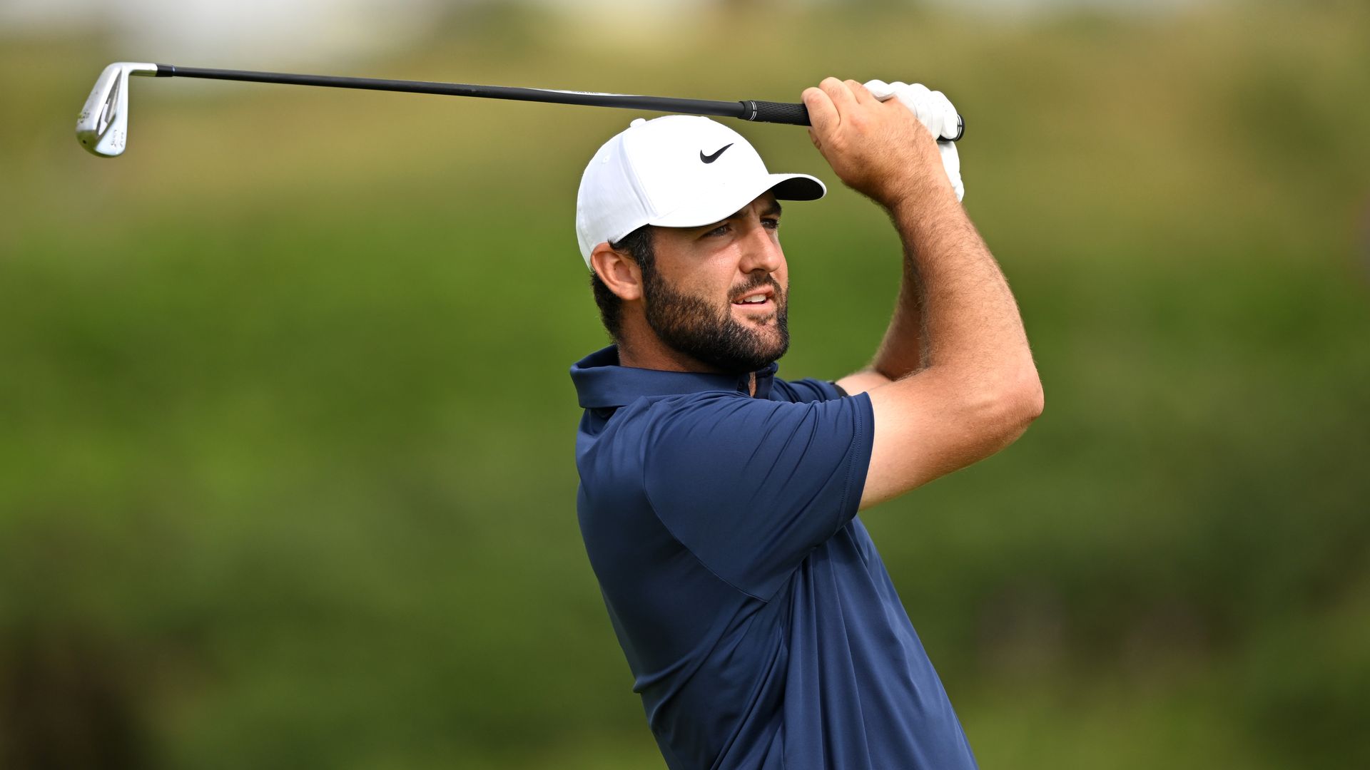 A male golfer in a white cap with a black Nike swoosh, wearing a blue polo, swings a golf club through his backswing with a determined expression; green, blurred course in background.