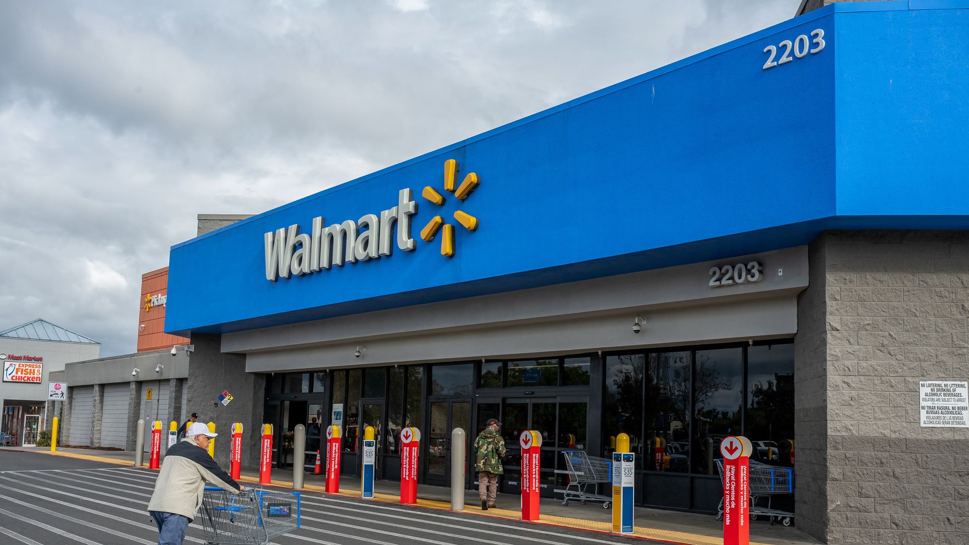 Outside view of a Walmart store with a large blue sign, store number 2203, people using shopping carts, and a cloudy sky overhead.