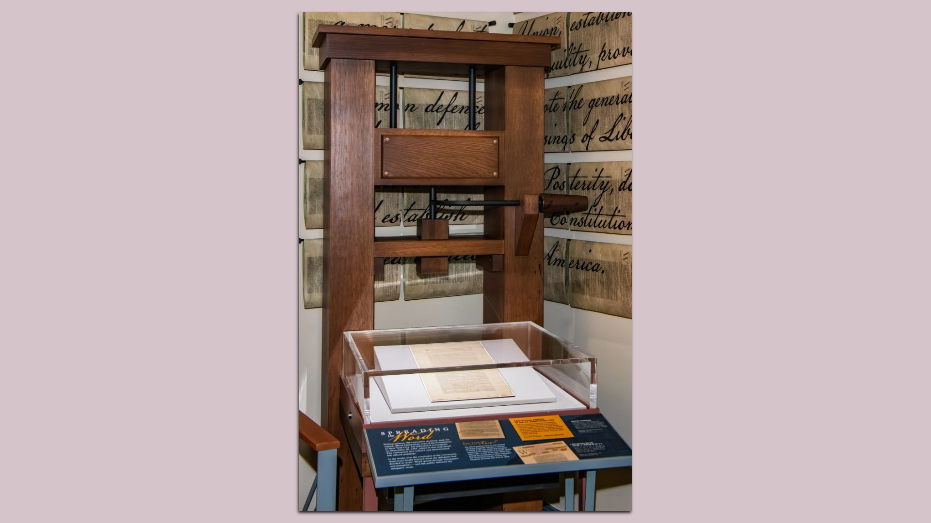 Wooden replica of an old printing press with a protected historic document on display beneath it, surrounded by vintage paper texts on the walls.