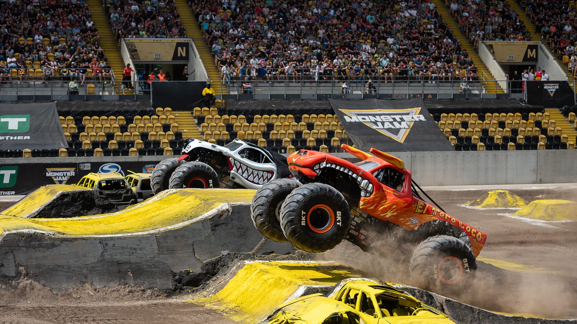 A photo of two monster trucks going over jumps in an arena.