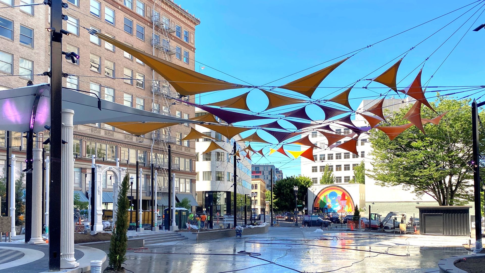 Urban plaza under orange and brown fabric shade sails stretched between black poles; a brick building with windows on the left, blue sky overhead, and a rainbow circular mural in the background.