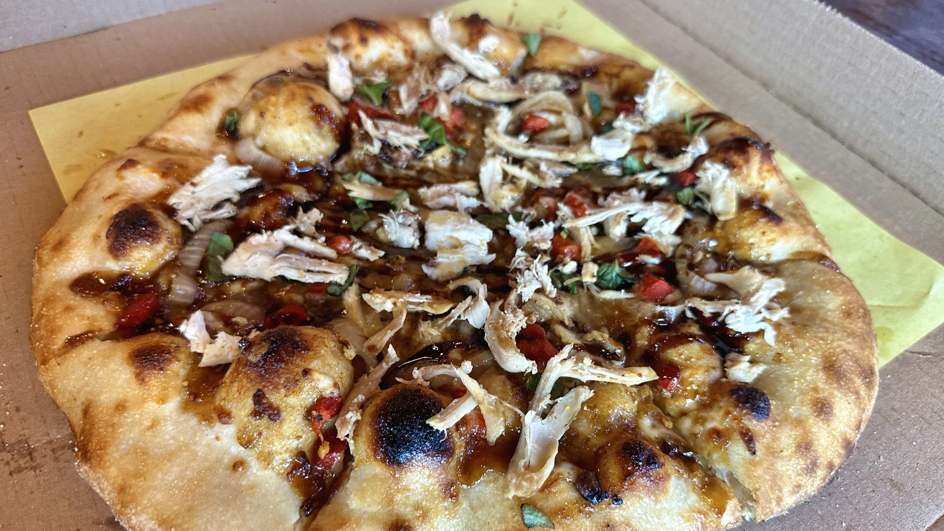 Pizza in a takeout box with a yellow liner; thick crust with char, topped with shredded chicken, onions, red peppers, and green herbs in a smoky BBQ sauce; several slices eaten.