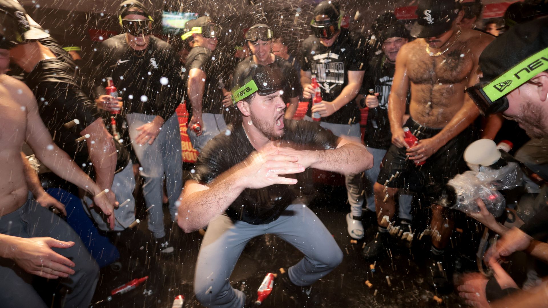 Group of men wearing goggles and headlamps at a crowded party, some shirtless, dancing as white confetti falls around them and red bottles are in their hands.