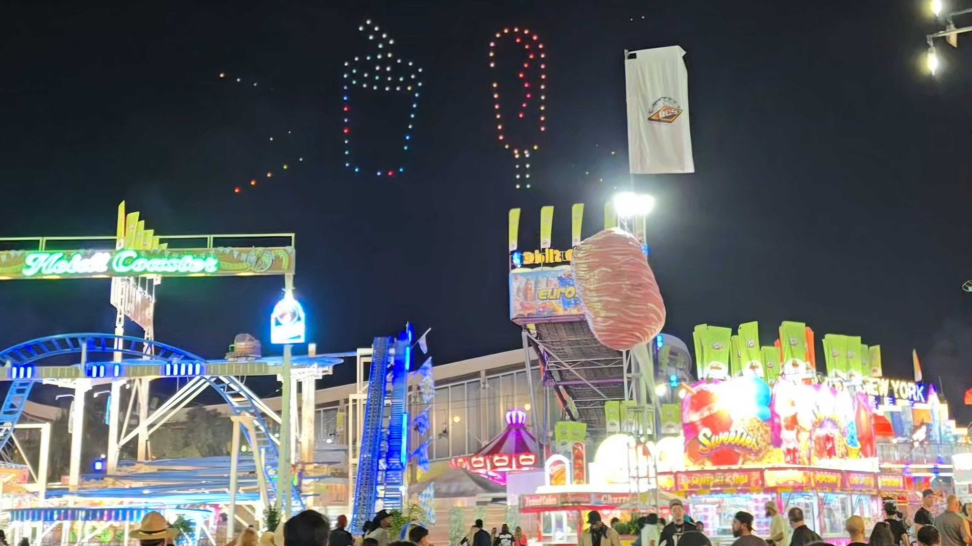 Night carnival with colorful LED light sculptures in the sky: a glowing circular ring, a tall multicolor candy-cane shape, and other dotted silhouettes above busy food stalls and a crowd.