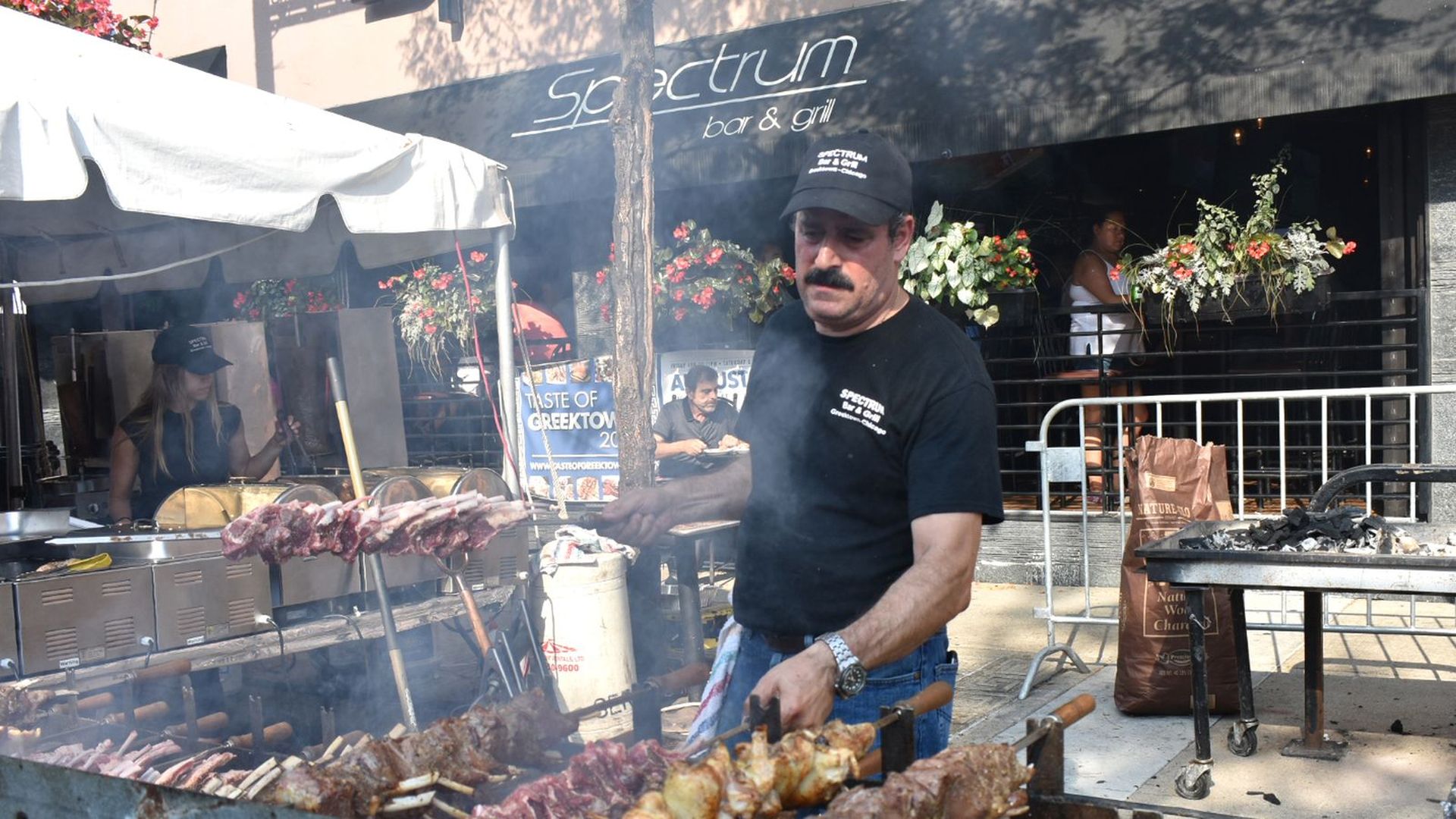 A photo showing a man outside cooking large kabobs over a smoking grill. He's in front of the Spectrum Bar & Grill.