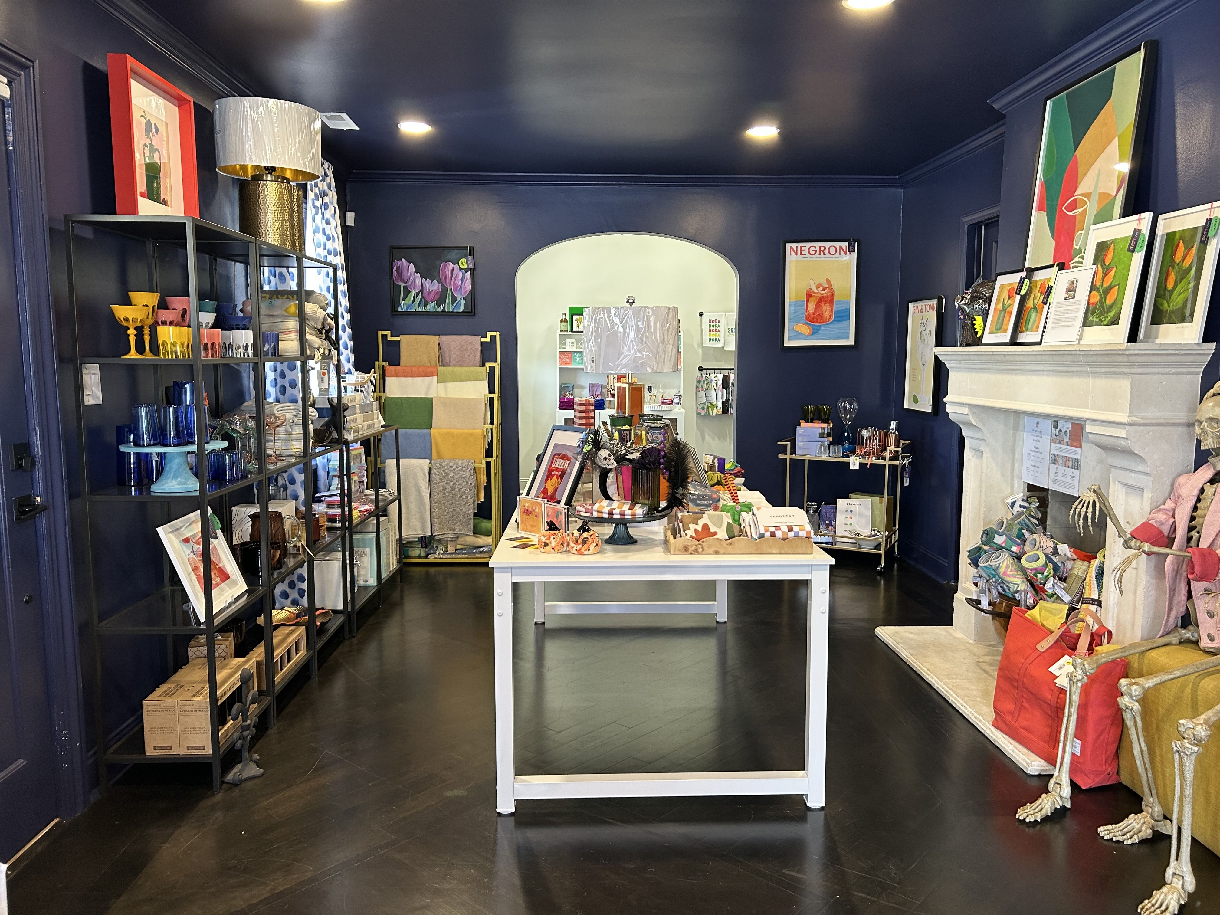 Boutique shop interior with dark blue walls and ceiling, white table displaying colorful items, black shelves on left with glassware and decor, fireplace on right with art and skeleton models.