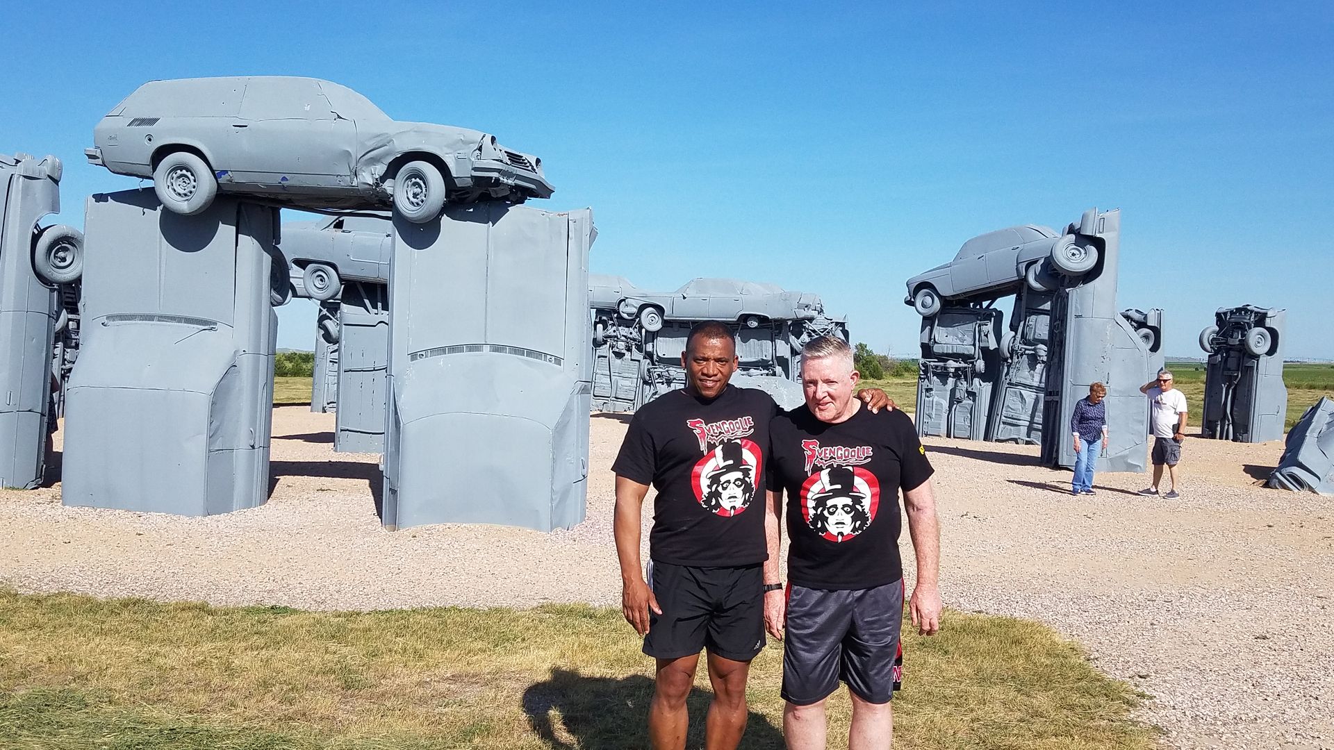 Two men standing in front of "Carhenge," a Stonehenge-style statue of cars on plinths.
