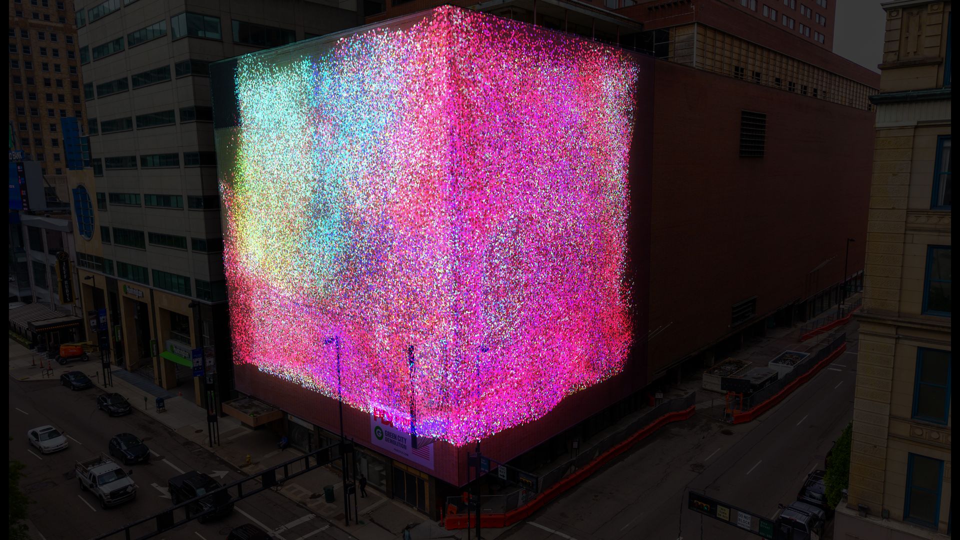 Urban street scene featuring a tall building wrapped in a massive LED screen with multicolored lights, creating a rainbow gradient across the façade, beside other city buildings and parked cars.
