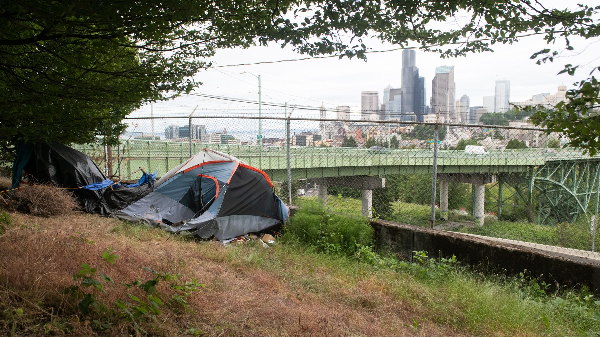 A tent sits on the edge of an embankment near a bridge and other roads with the Seattle skyline in the far background.