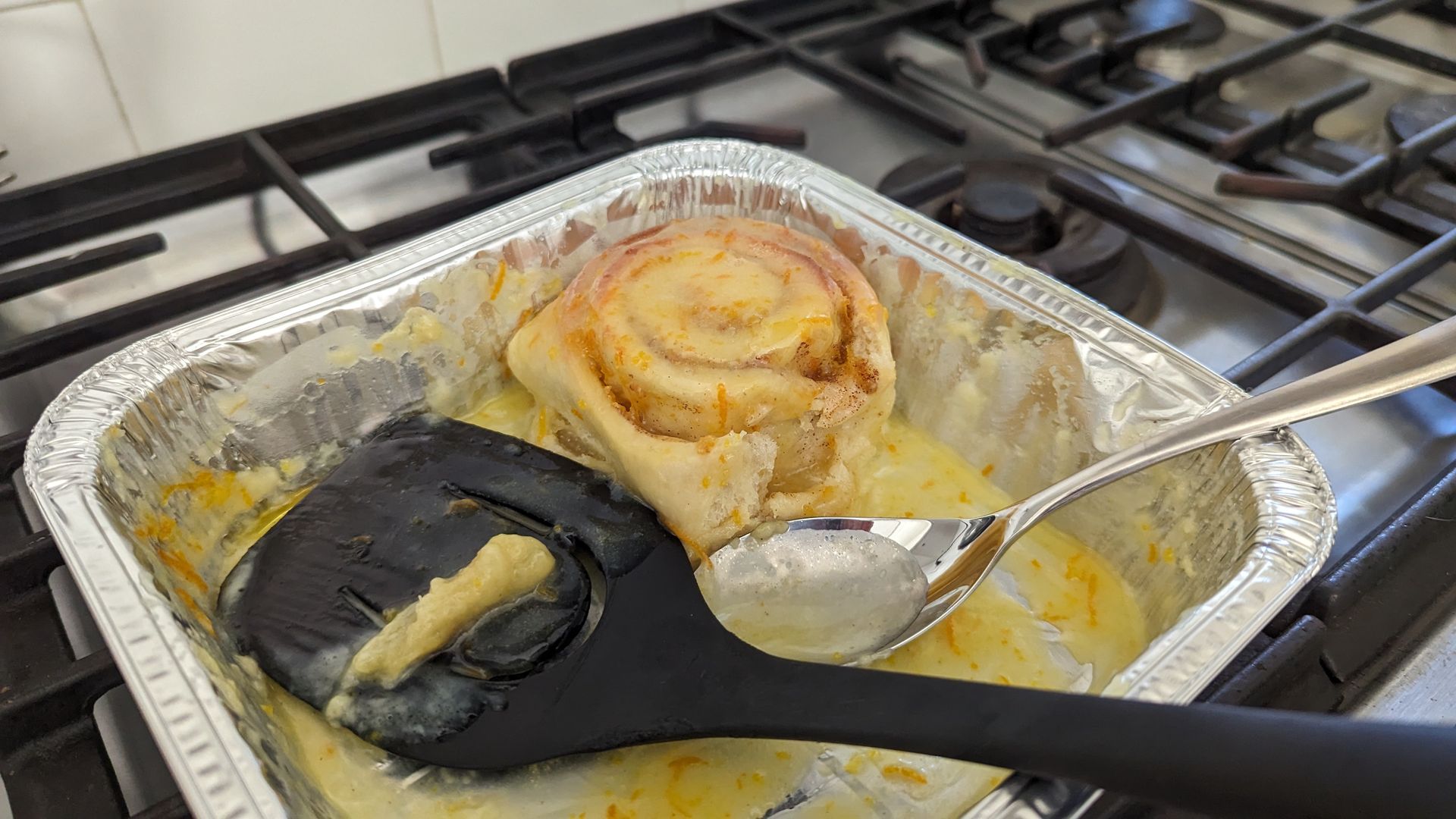 A hot roll in a pan with frosting, a spatula and a spoon.