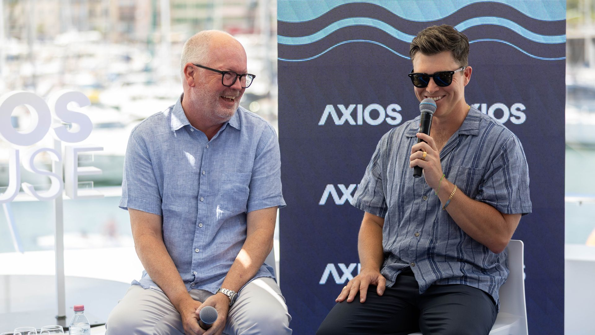 Mark Marshall chairman of global advertising and partnerships at NBCUniversal and SNL star Colin Jost sit on a panel in front of a blue Axios banner with wave patterns; Marshall on the left in light blue shirt and glasses smiles, Jost in a striped shirt and sunglasses speaks into a microphone.
