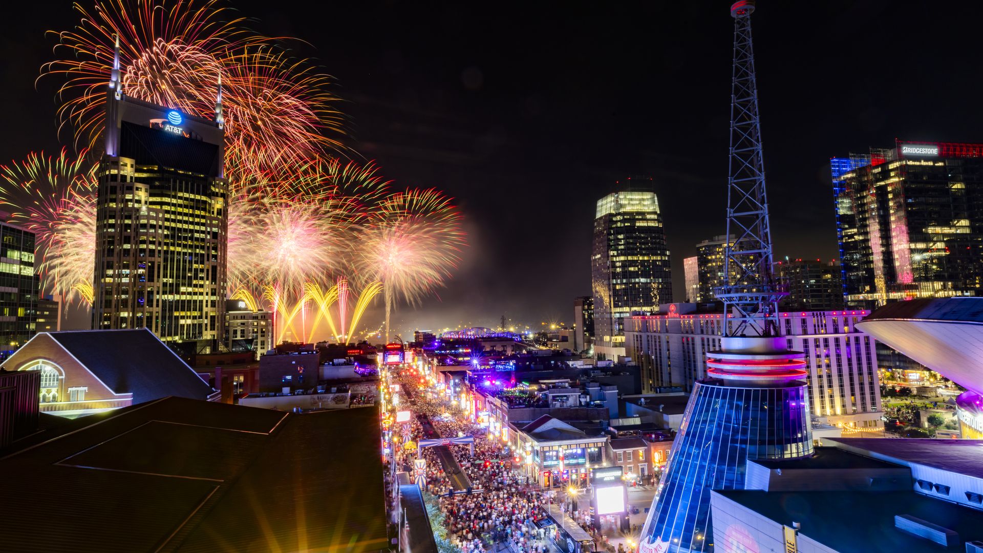 Nighttime city skyline as colorful fireworks explode over the AT&T building, a tall lattice tower on the right, and a crowded street festival glowing with neon lights.