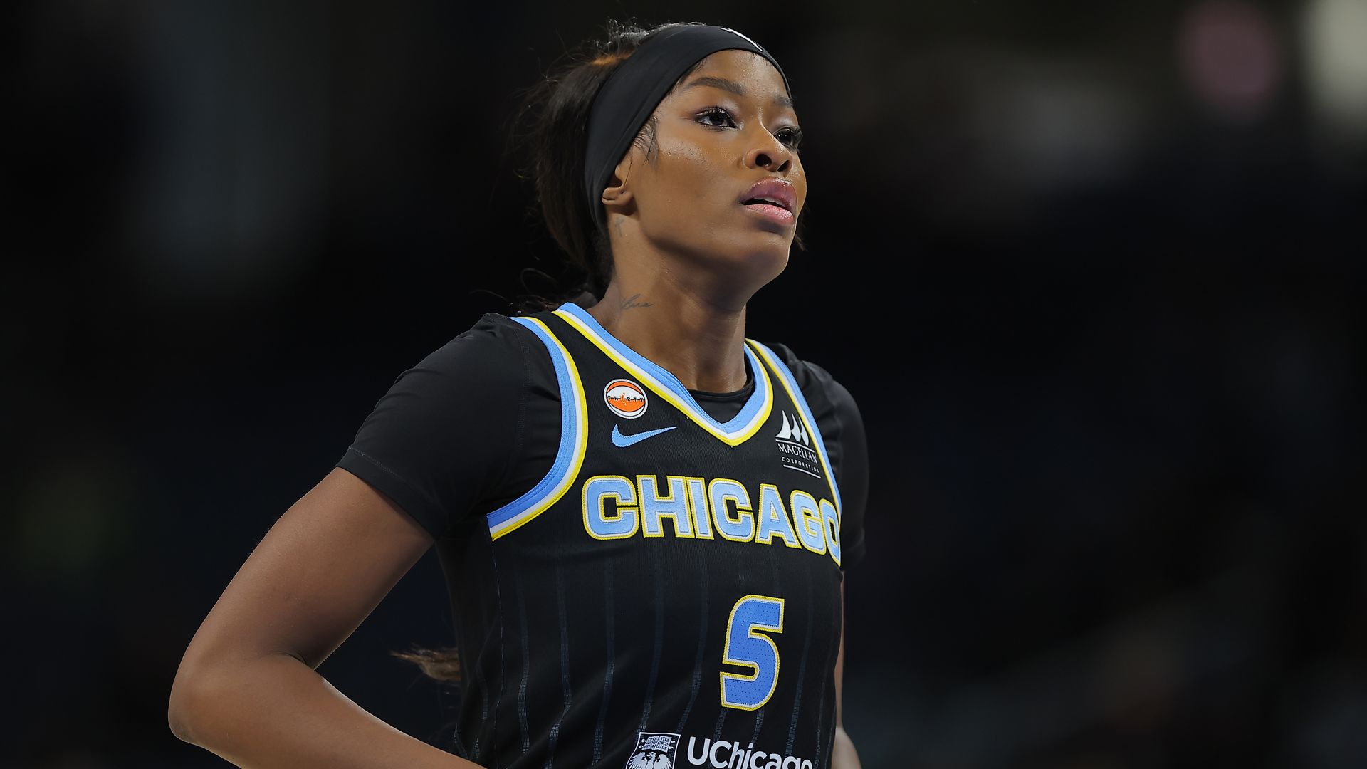 Close-up of a Black female basketball player wearing a dark Chicago jersey with blue and yellow trim and a black headband, looking upward with a blurred arena background.