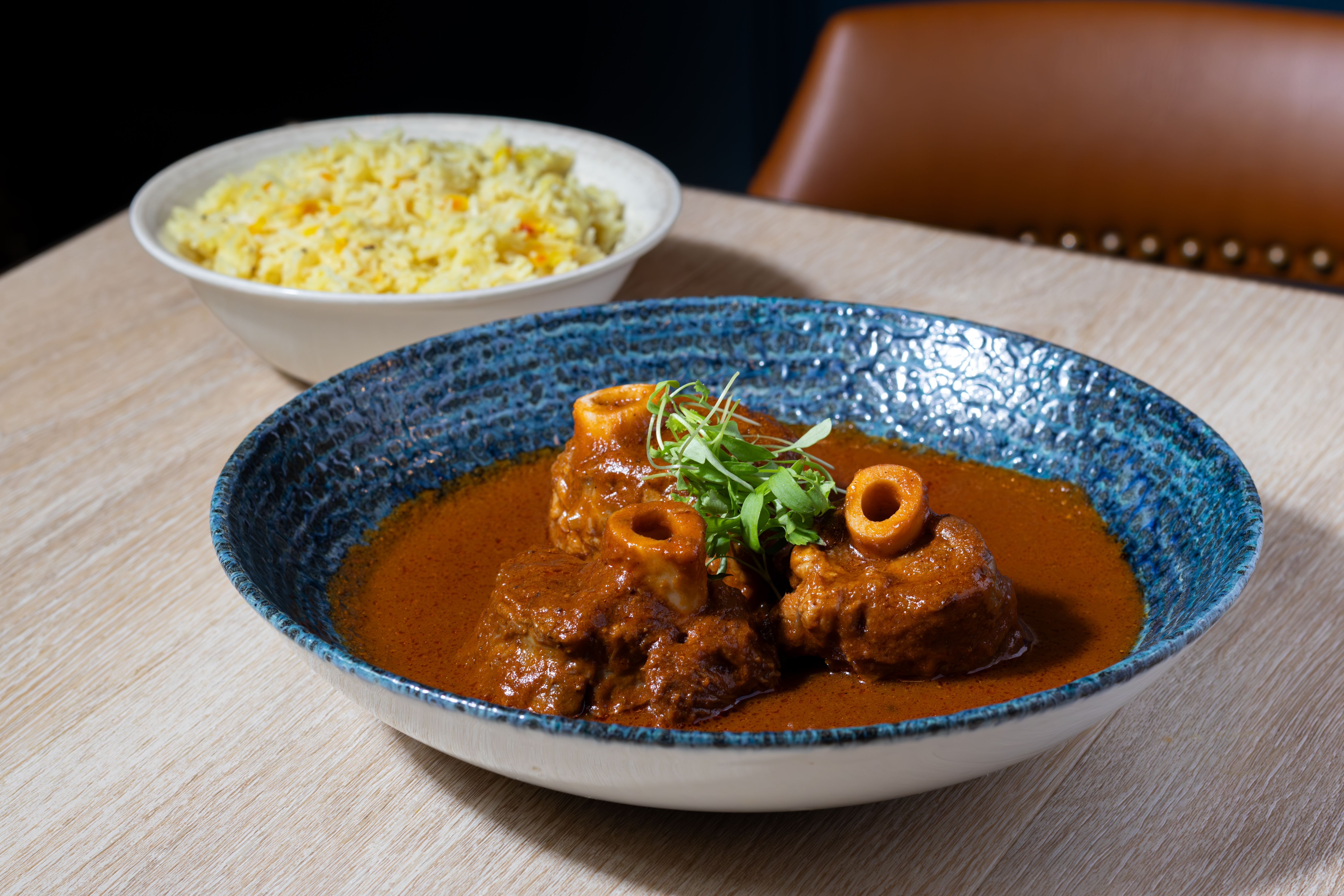 Blue textured bowl of orange curry with meat chunks and microgreens on a wooden table; behind it is a white bowl of yellow rice, and a brown chair back against a dark background.
