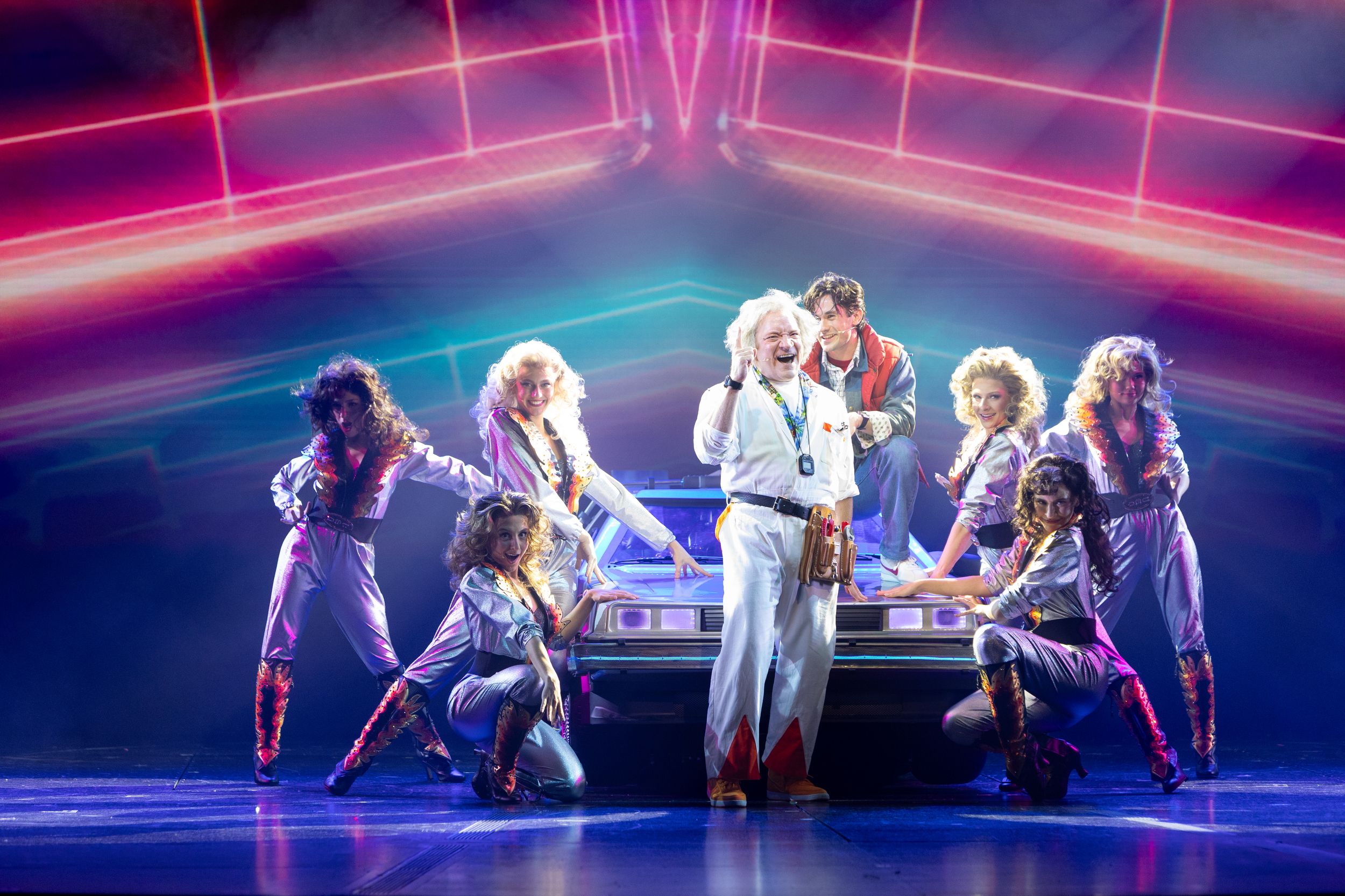Stage performance with seven people around a silver car under pink and blue neon lights. One man in white jumpsuit smiles while others wear silver outfits with flames, posing energetically.
