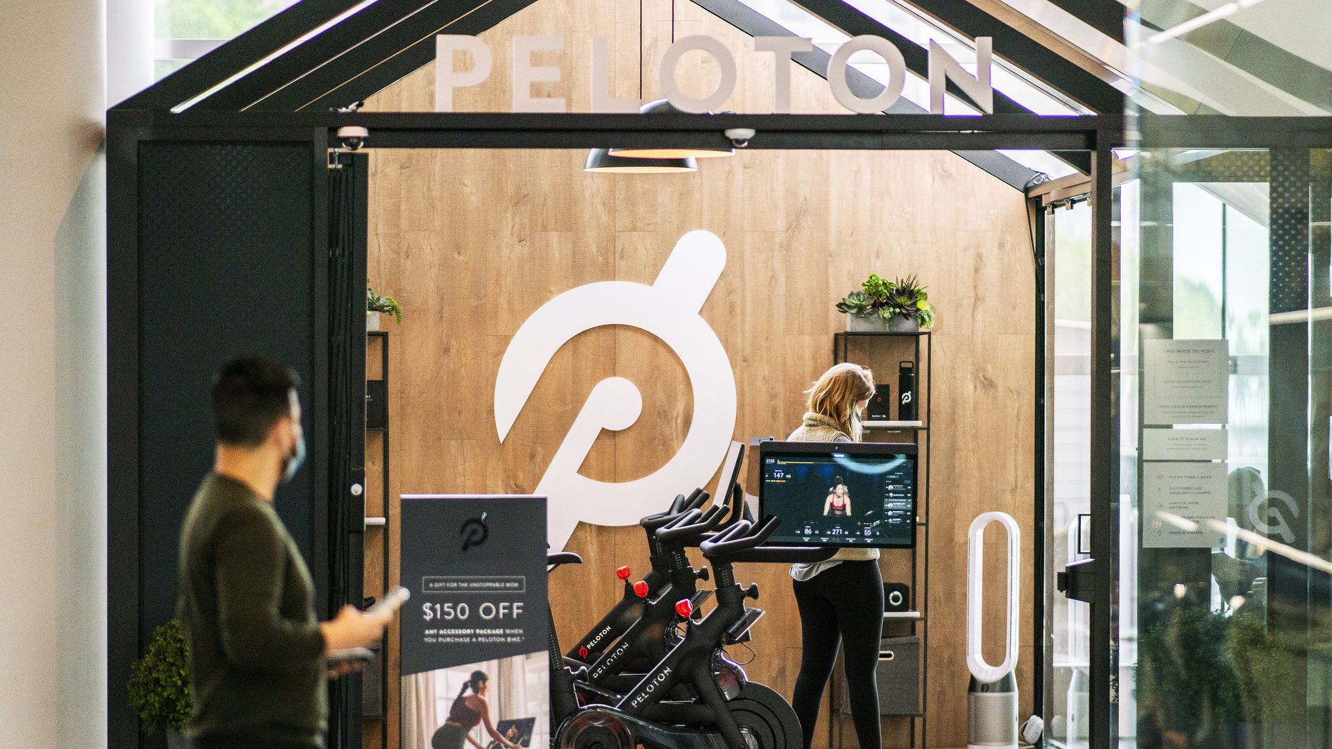 A man walks in front of a Peloton store with two Peloton bikes and a woman inside.