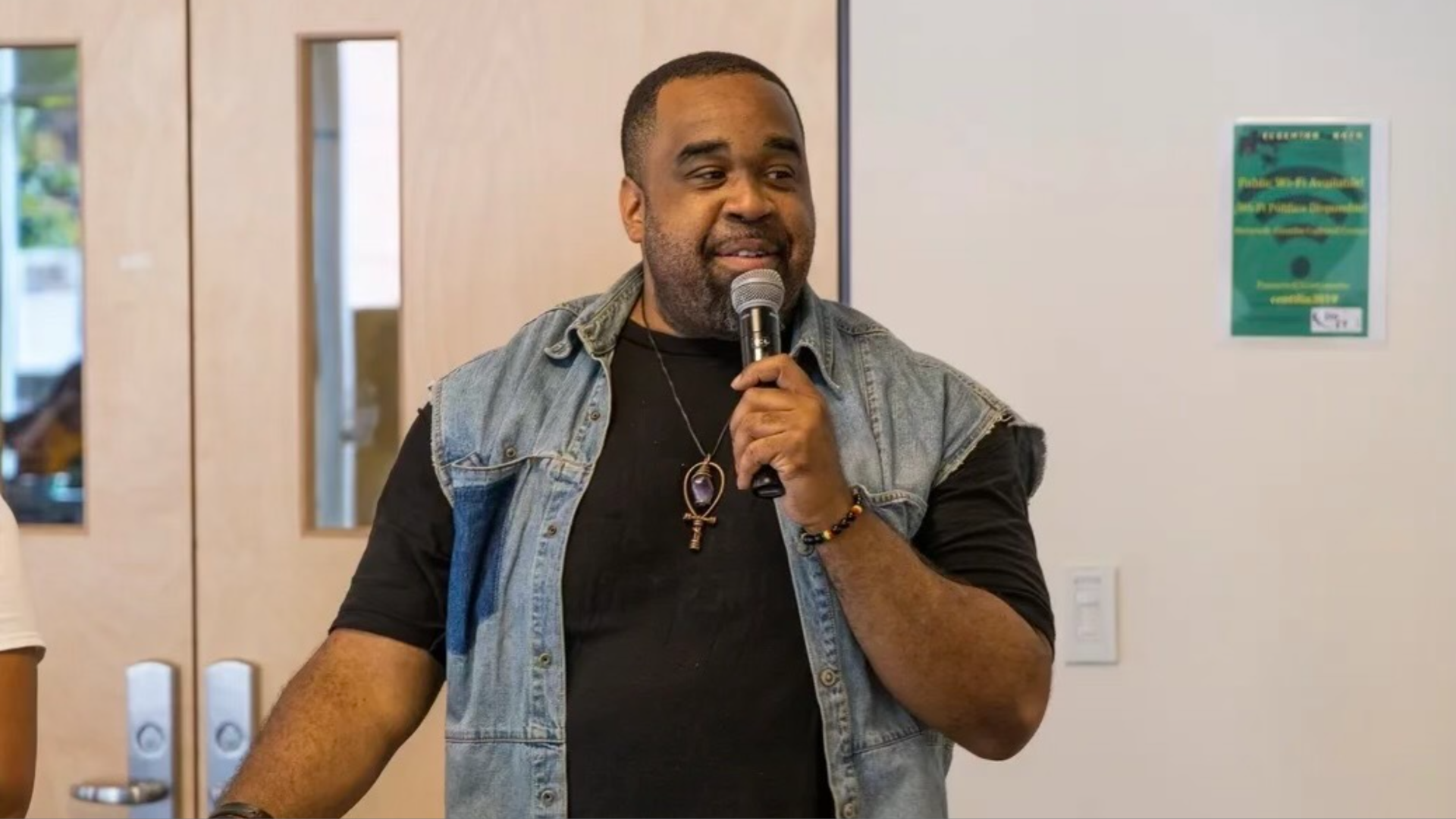 Jeffrey Cheatham speaks into a microphone in a room with neutral tones. He is wearing a denim vest and a black T-shirt. 