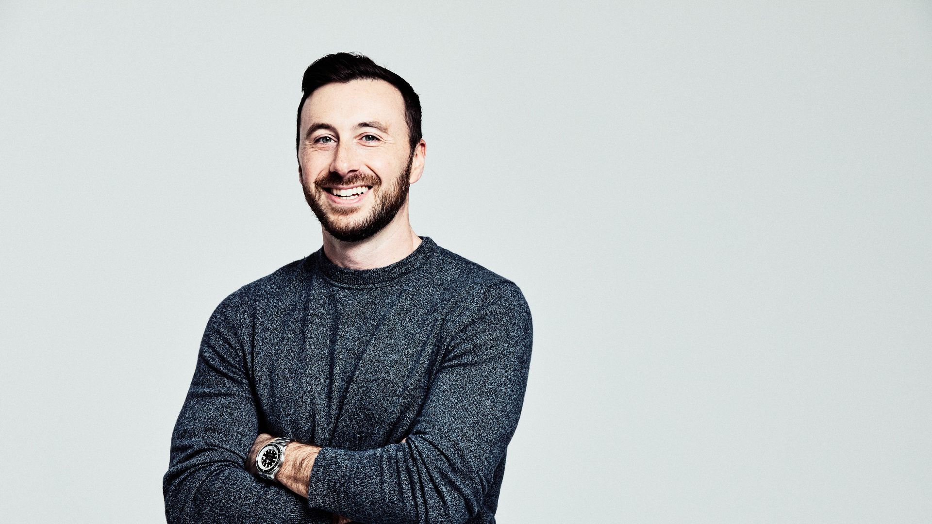 Portrait of a smiling man with short dark hair and a trimmed beard, wearing a speckled dark blue sweater and a wristwatch, arms crossed against a light gray background.