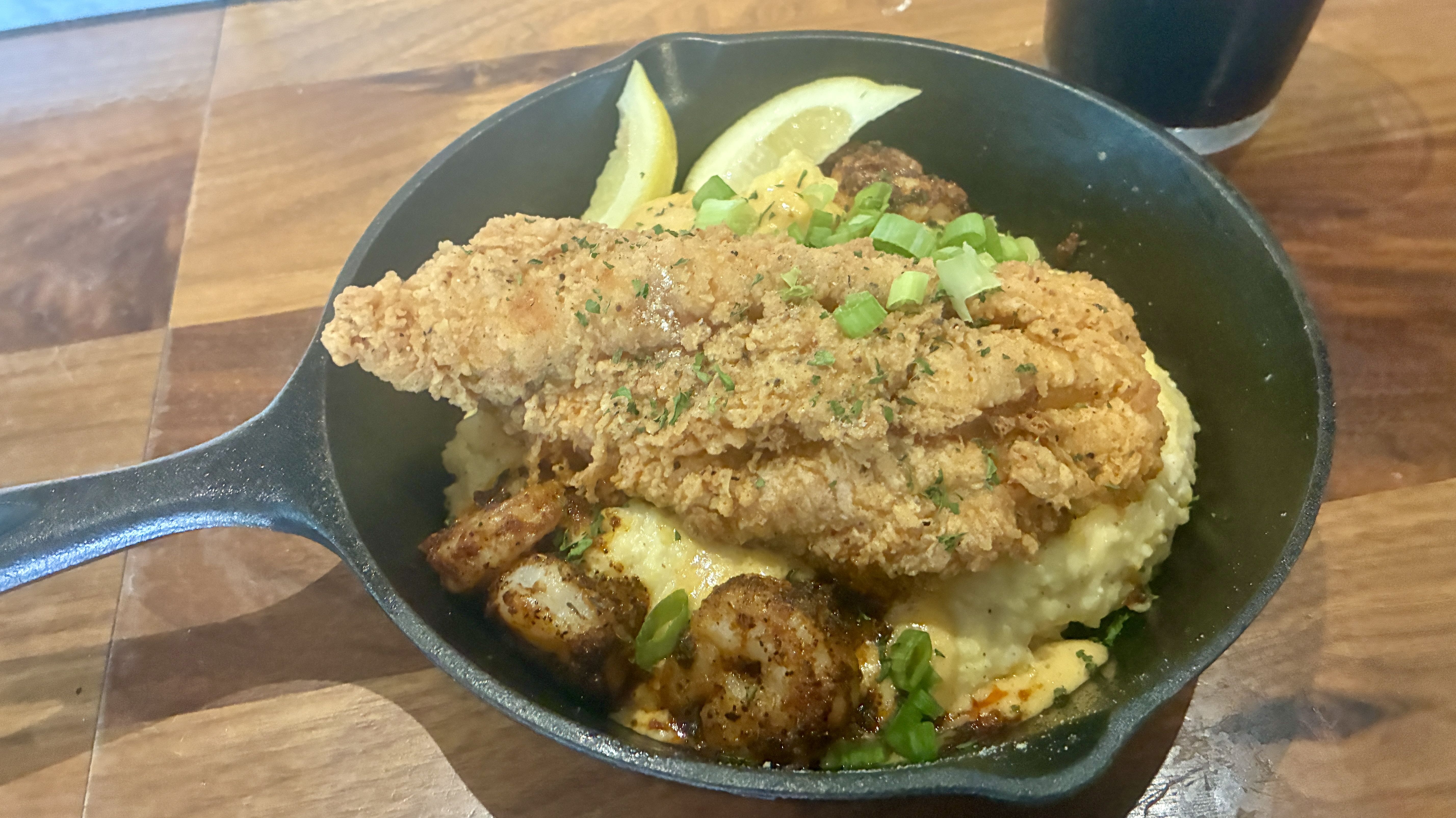 Crispy fried chicken strips atop creamy mashed potatoes and gravy in a black skillet, garnished with chopped green onions and lemon wedges on a wooden table.