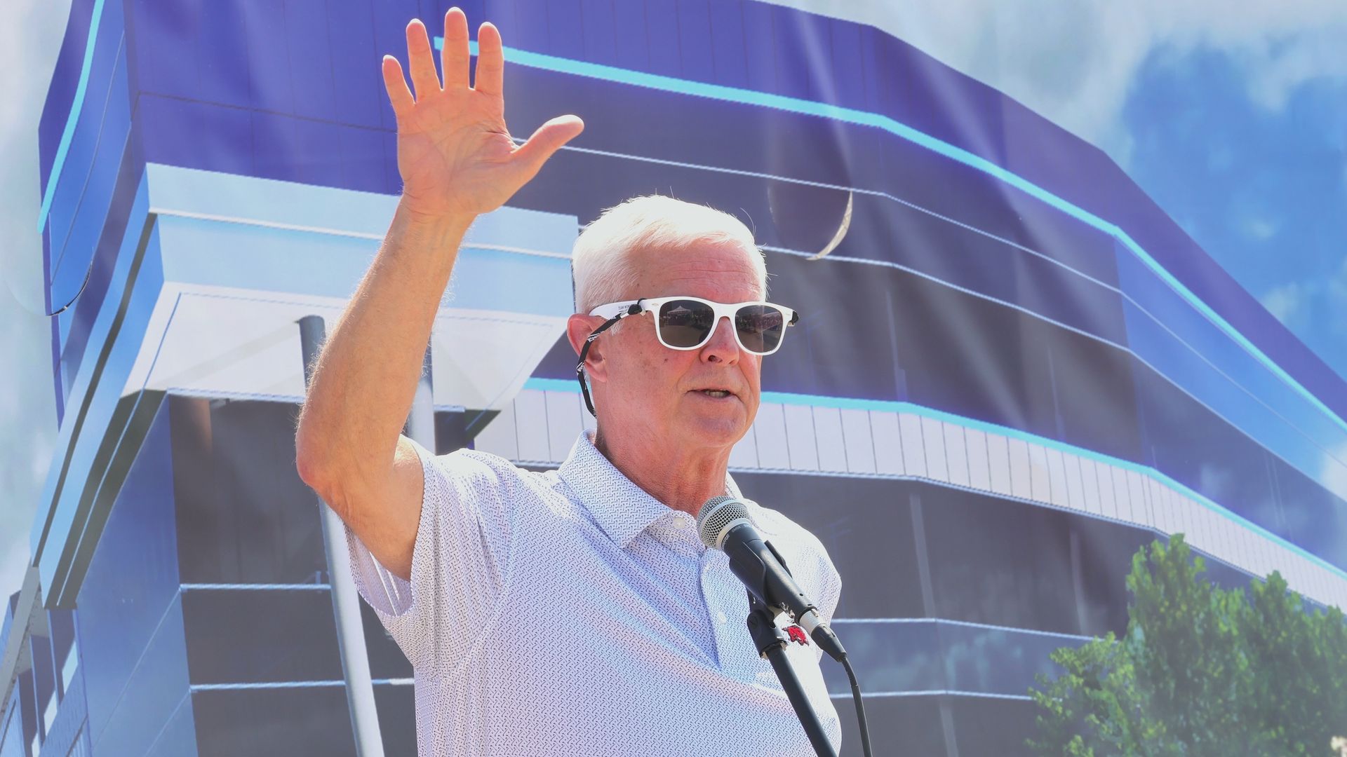 U.S. Rep. Steve Womack speaks at a podium outdoors, raising his hand in front of a banner displaying a futuristic blue building.