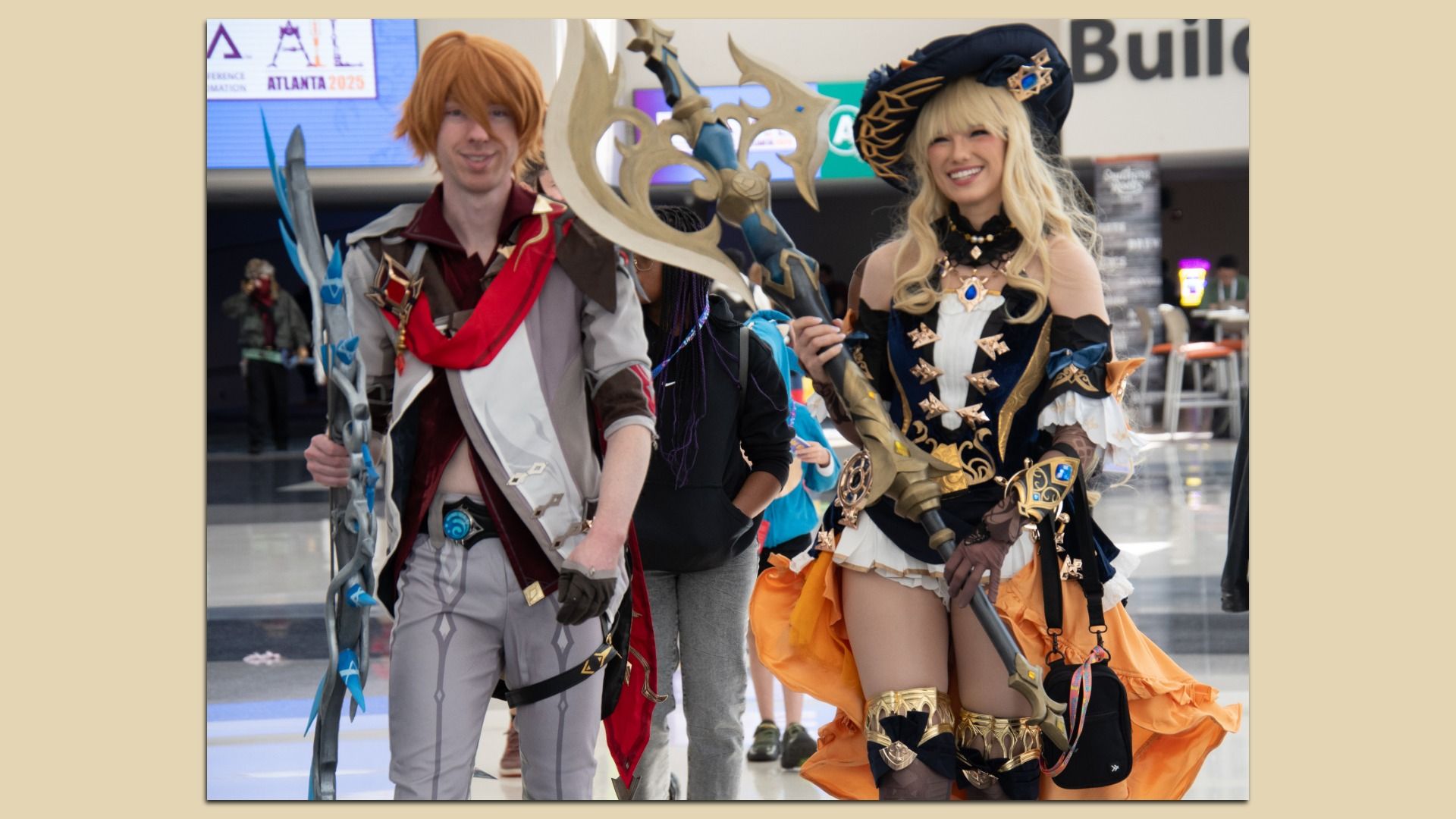 Two cosplayers pose at a convention: left, a man in grey-red armor holding a blue crystal staff; right, a smiling blonde woman in a black-gold corset costume with a large hat.