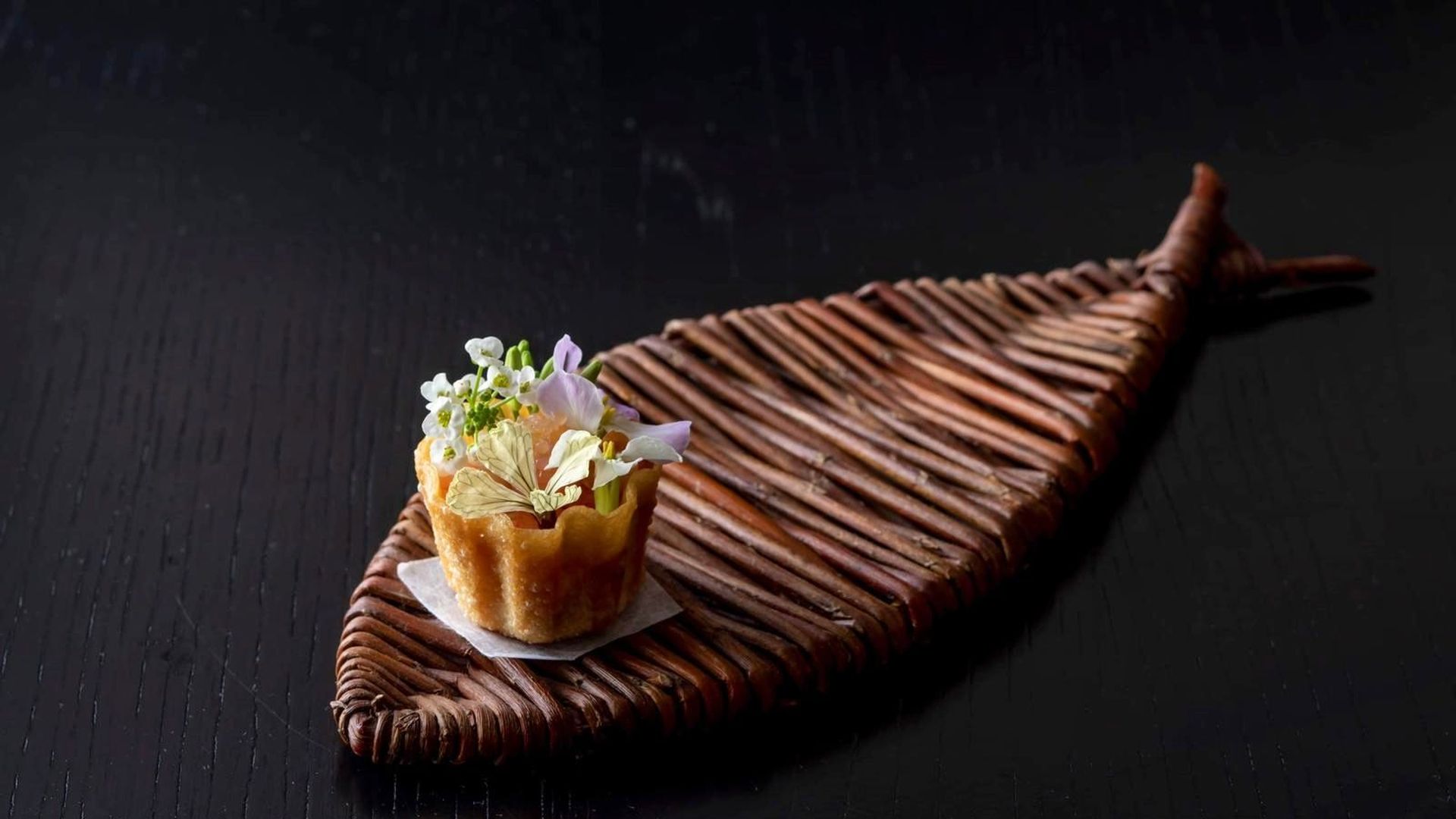 A crispy wonton-like appetizer, filled with edible flowers, served on a leaf-shaped wood woven tray.