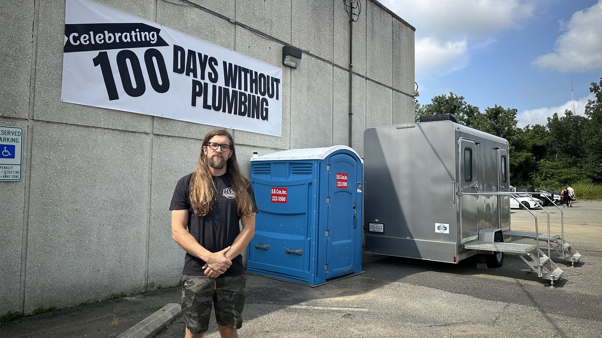 A man stands in front of a sign that says 100 days without plumbing