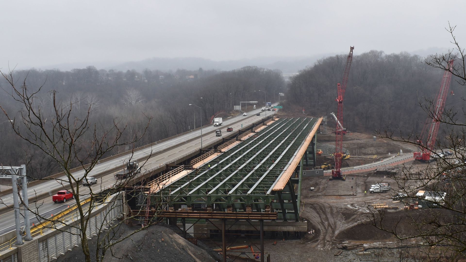 Construction site of a large green steel bridge parallel to an existing highway with vehicles, cranes, bare trees, and foggy hills in the background under a gray sky.