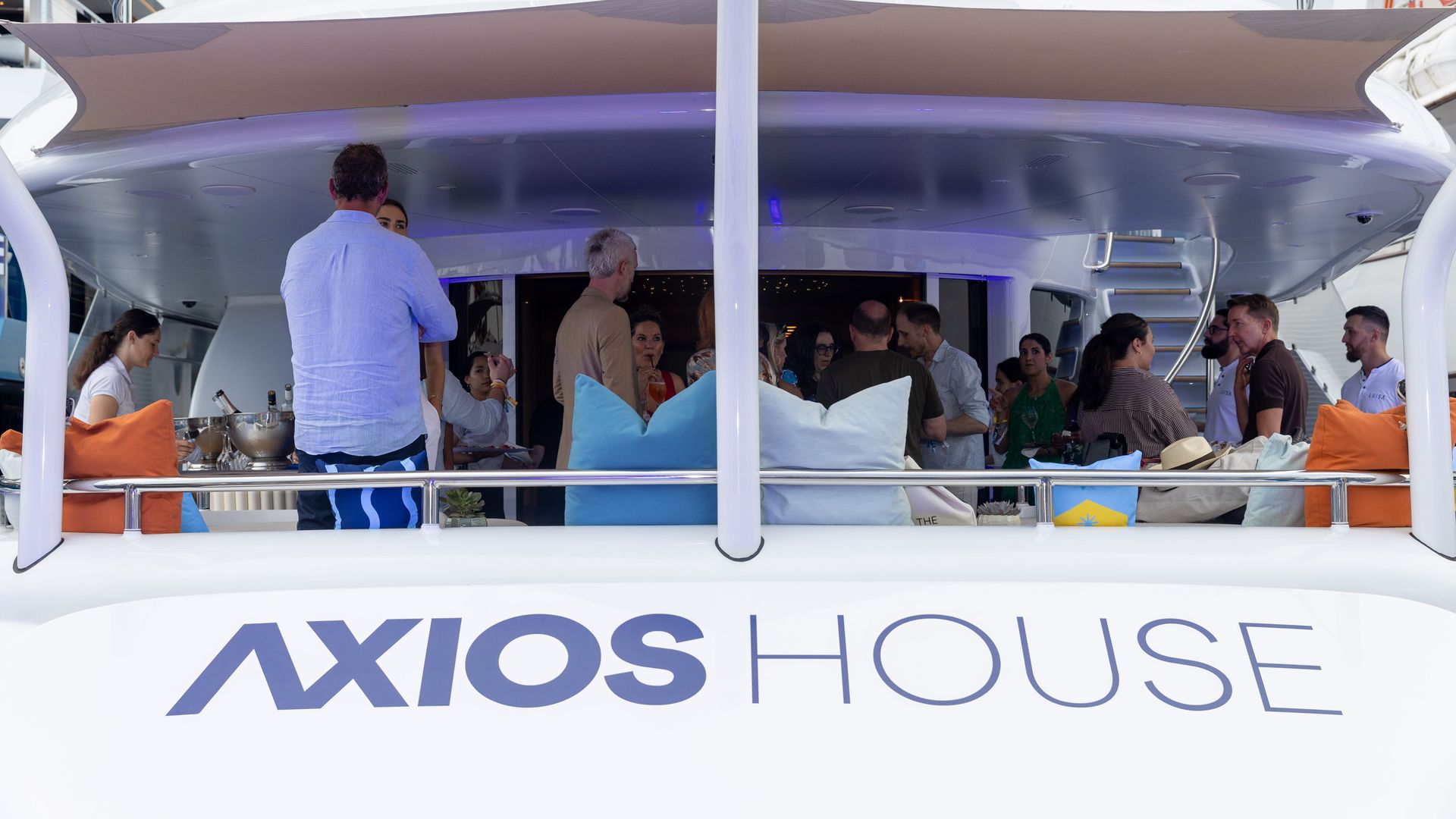People mingle on a large white luxury yacht with a beige shade sail and orange and blue cushions; guests stand on the open deck, while the hull shows Axios House.