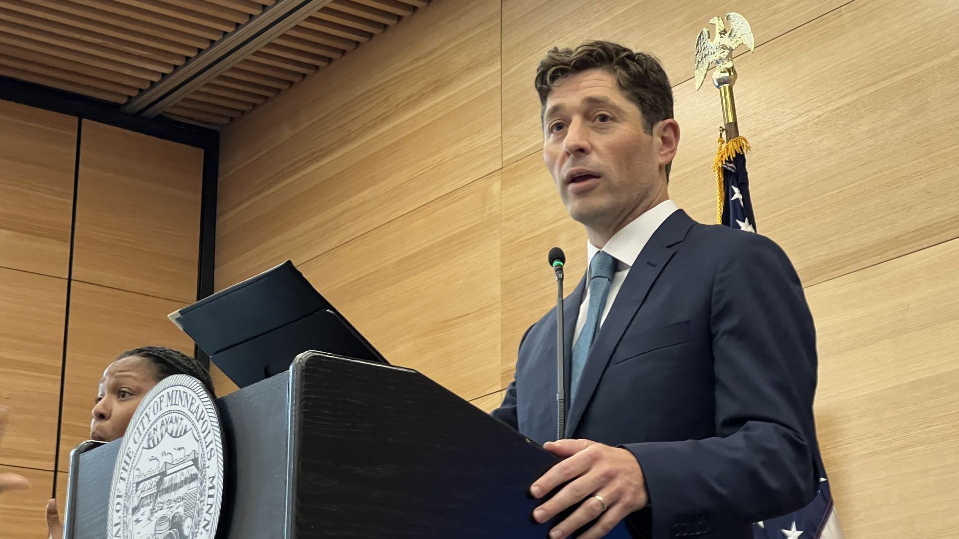 A man with light skin wearing a blue suit and a blue tie stands at a lectern flanked by an American flag in a wood-paneled room