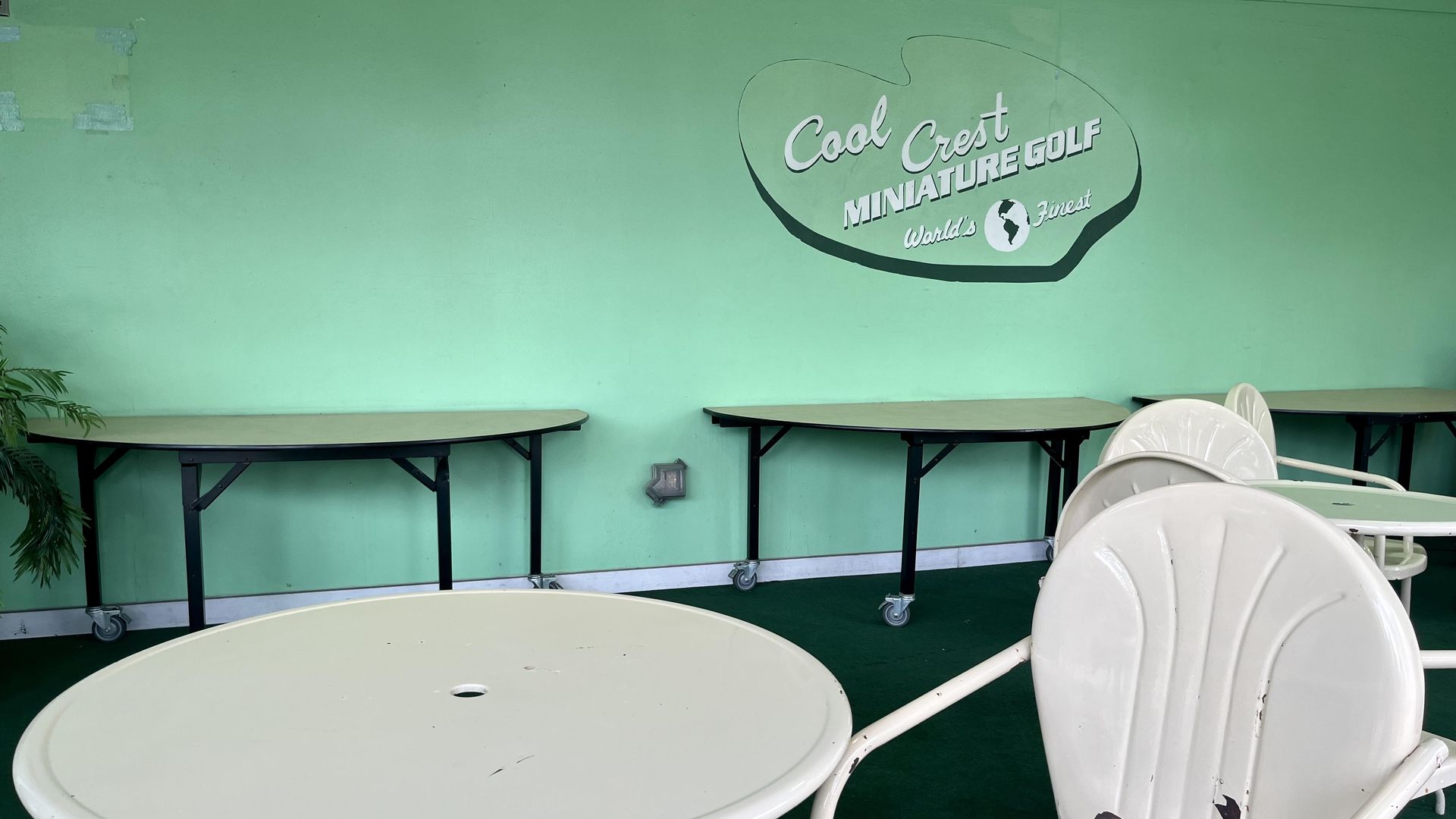 A seating area with mint green paint and white MCM tables with a painted sign for Cool Crest Mini Golf on the wall. 