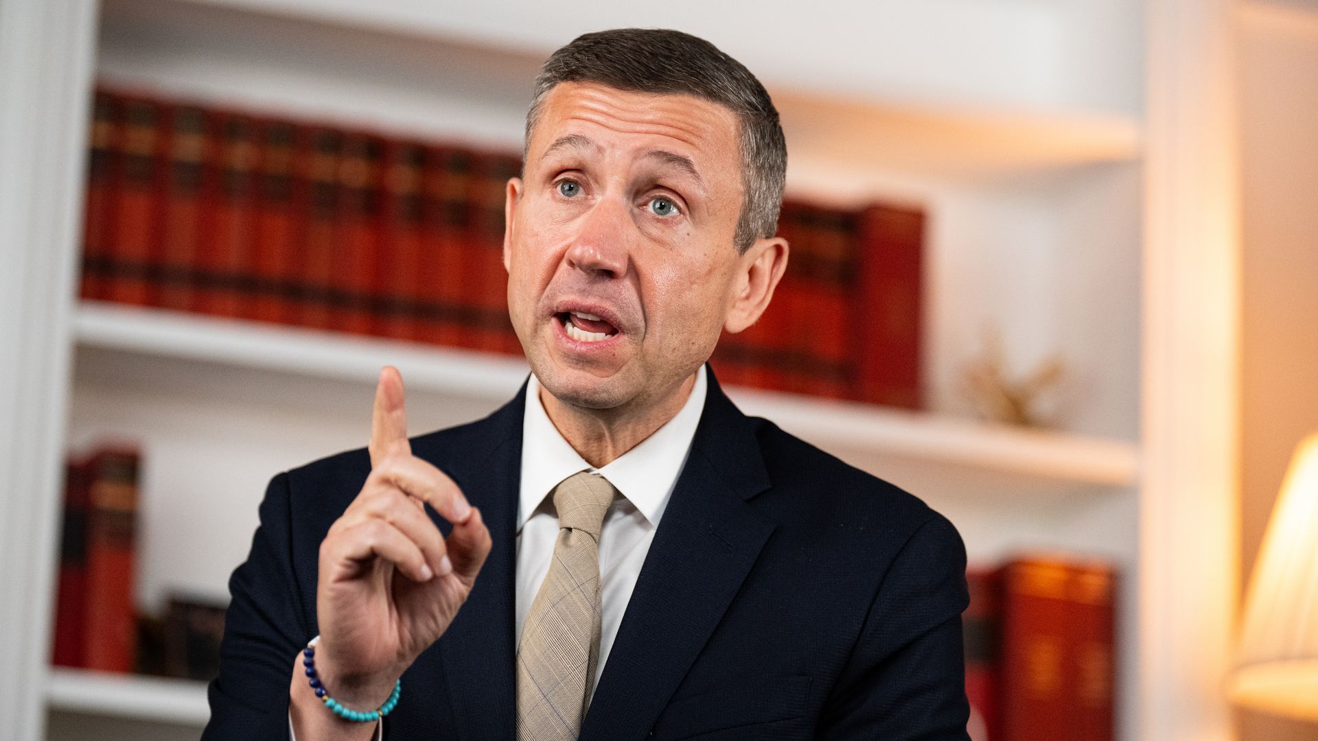 A man in a dark suit and beige tie speaks, gesturing with his index finger. He wears a turquoise bracelet; red bookshelves and warm, blurred lighting fill the background.