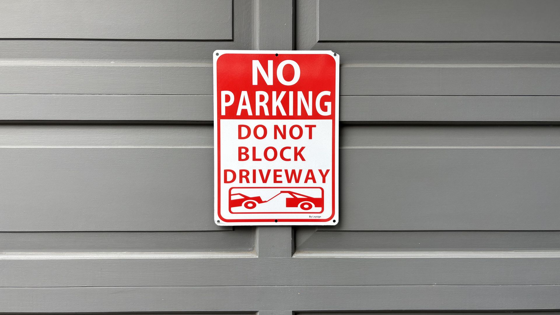 Red and white 'NO PARKING DO NOT BLOCK DRIVEWAY' sign on a gray garage door, with a small tow-truck graphic at the bottom. The sign is bordered in white and mounted with screws.