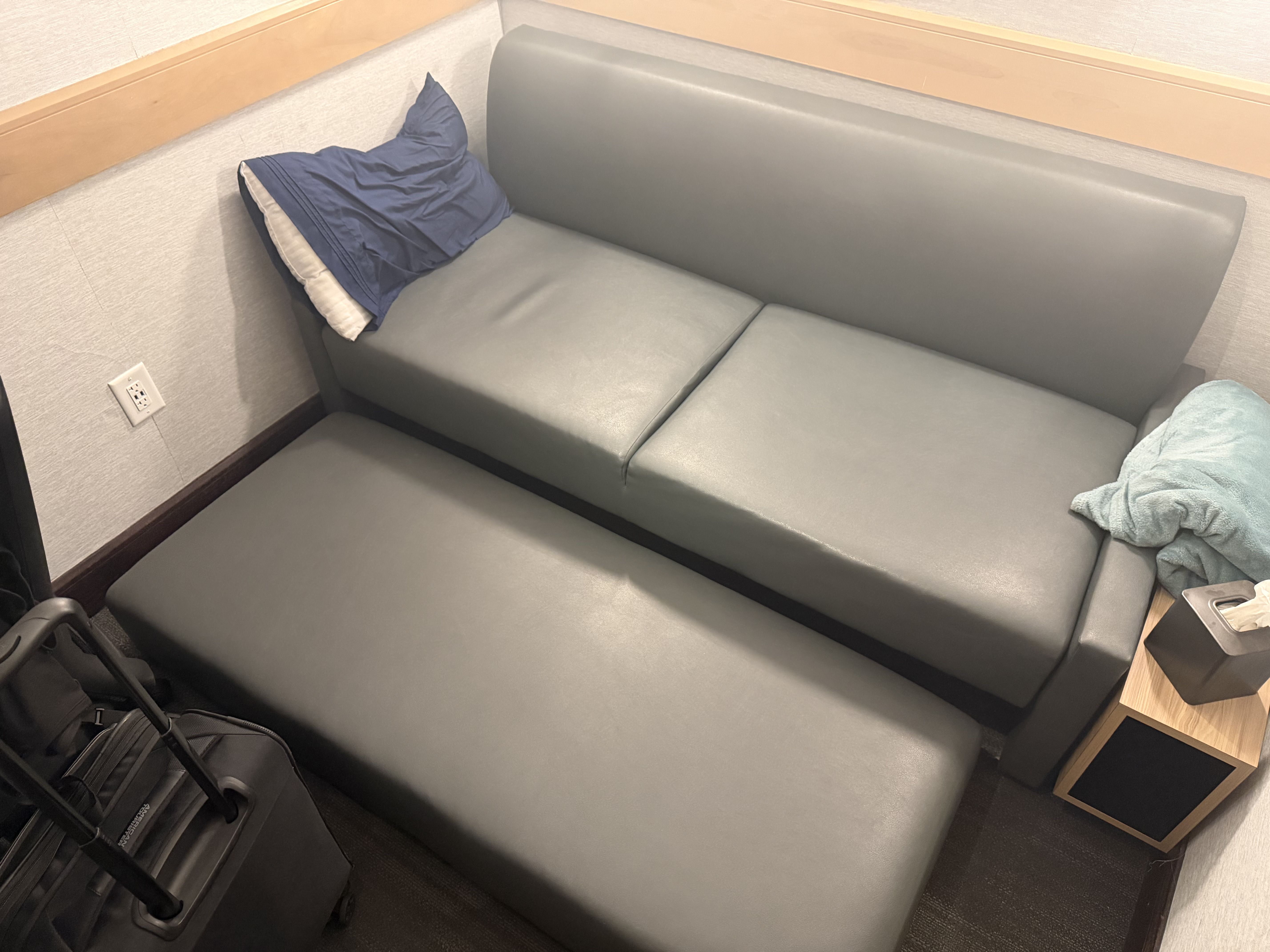 Small booth with a gray-blue sofa, a navy cushion in the corner, a light blue blanket on the right, beige walls with wood trim, and a wall outlet on the left.