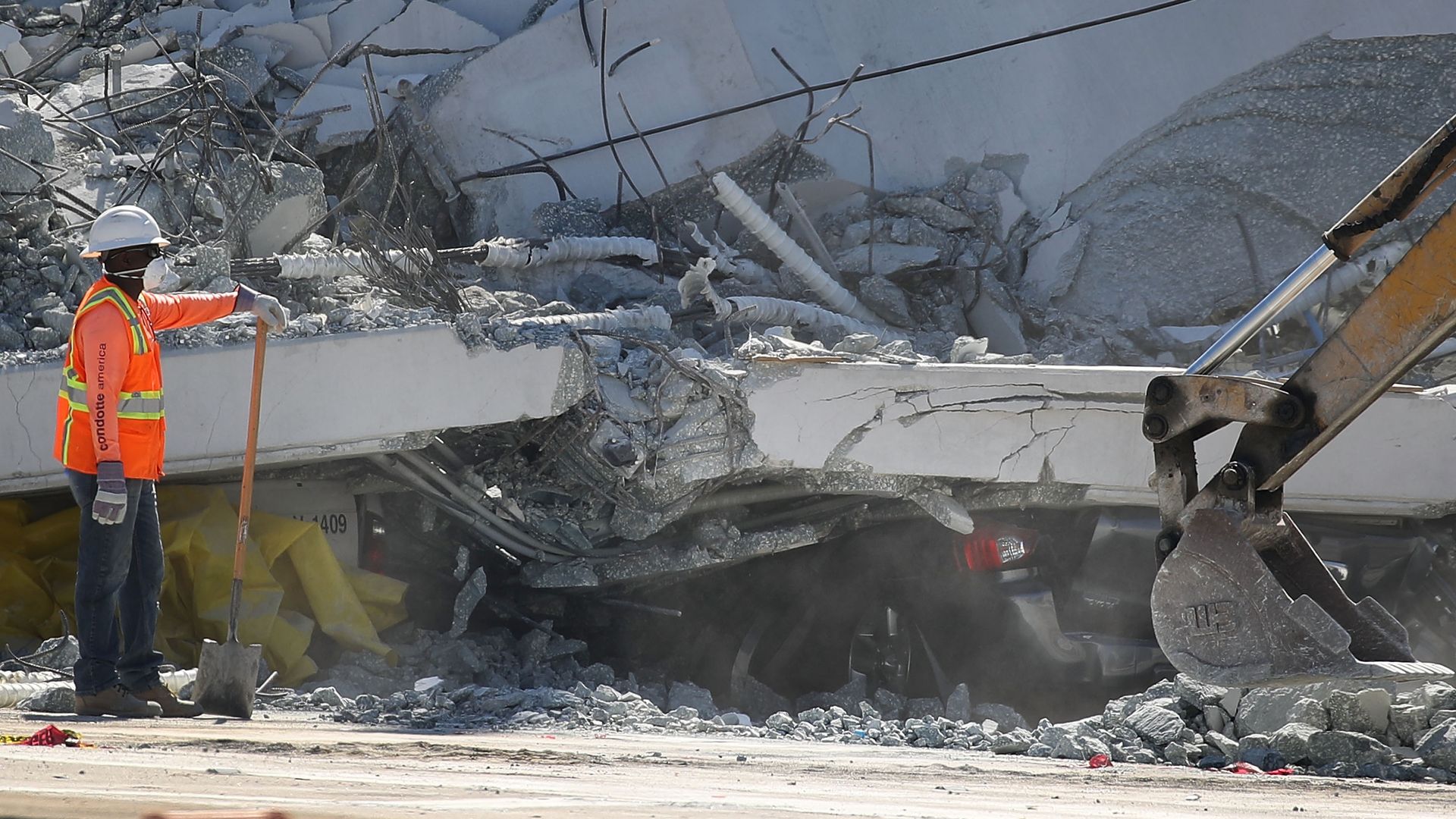 A crushed vehicle is seen near a worker as law enforcement and members of the National Transportation Safety Board investigate the scene where a pedestrian bridge collapsed
