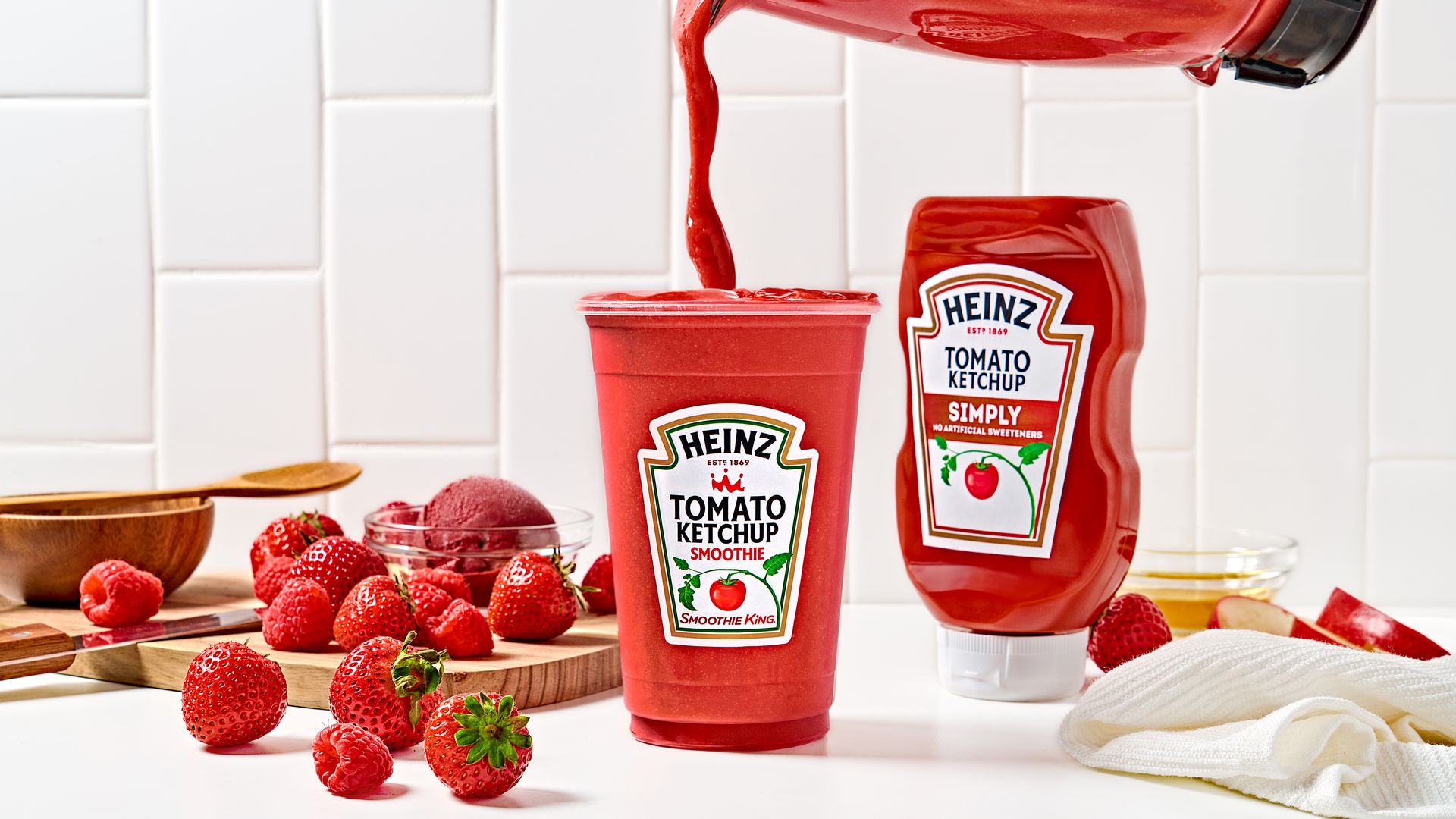 Heinz Tomato Ketchup Smoothie being poured into a cup, surrounded by fresh strawberries and raspberries, a bowl and spoon, and a Heinz Simply Ketchup bottle on a white kitchen counter.