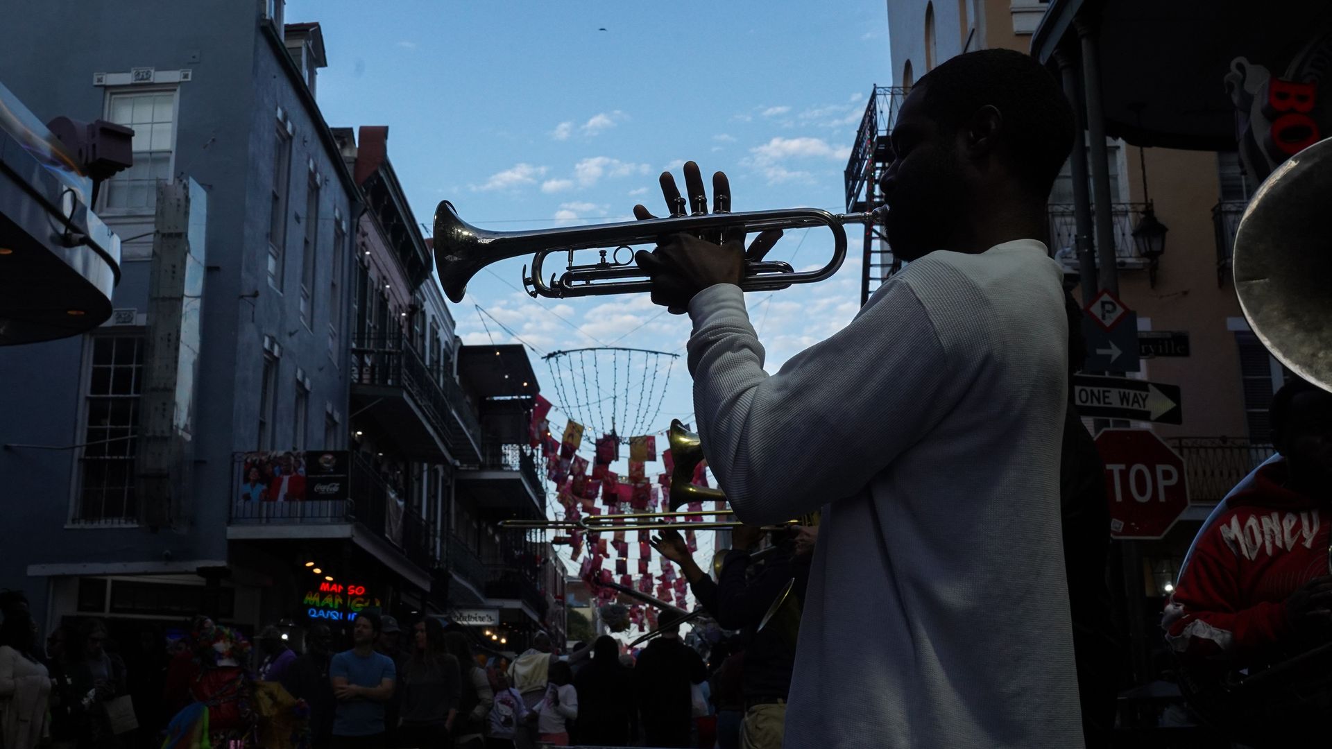 A man plays a trumpet on Bourbon Street. Storefronts and prayer flags strung up between buildings can be seen behind him against a blue sky.